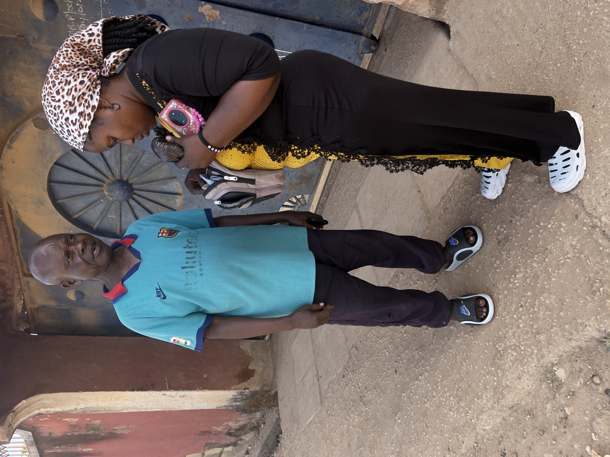 A medium shot features a Black adult man and a Black adult woman standing outside in Jos, Nigeria. The man wears a blue and red Barcelona football shirt, black trousers, and black and white Nike sandals. He looks toward the camera. The woman, standing on the opposite side, wears a black dress with yellow lace trim, a leopard-print head covering, and white slip-on shoes. She is looking down and fiddling with a small, dark object held in her hands. A pink camera bag is slung over her shoulder, and a beaded bracelet is visible on her wrist. The area outside is concrete. A large, dark metal door with decorative wheel-like spokes is behind the woman. A section of a brick wall is behind the man.