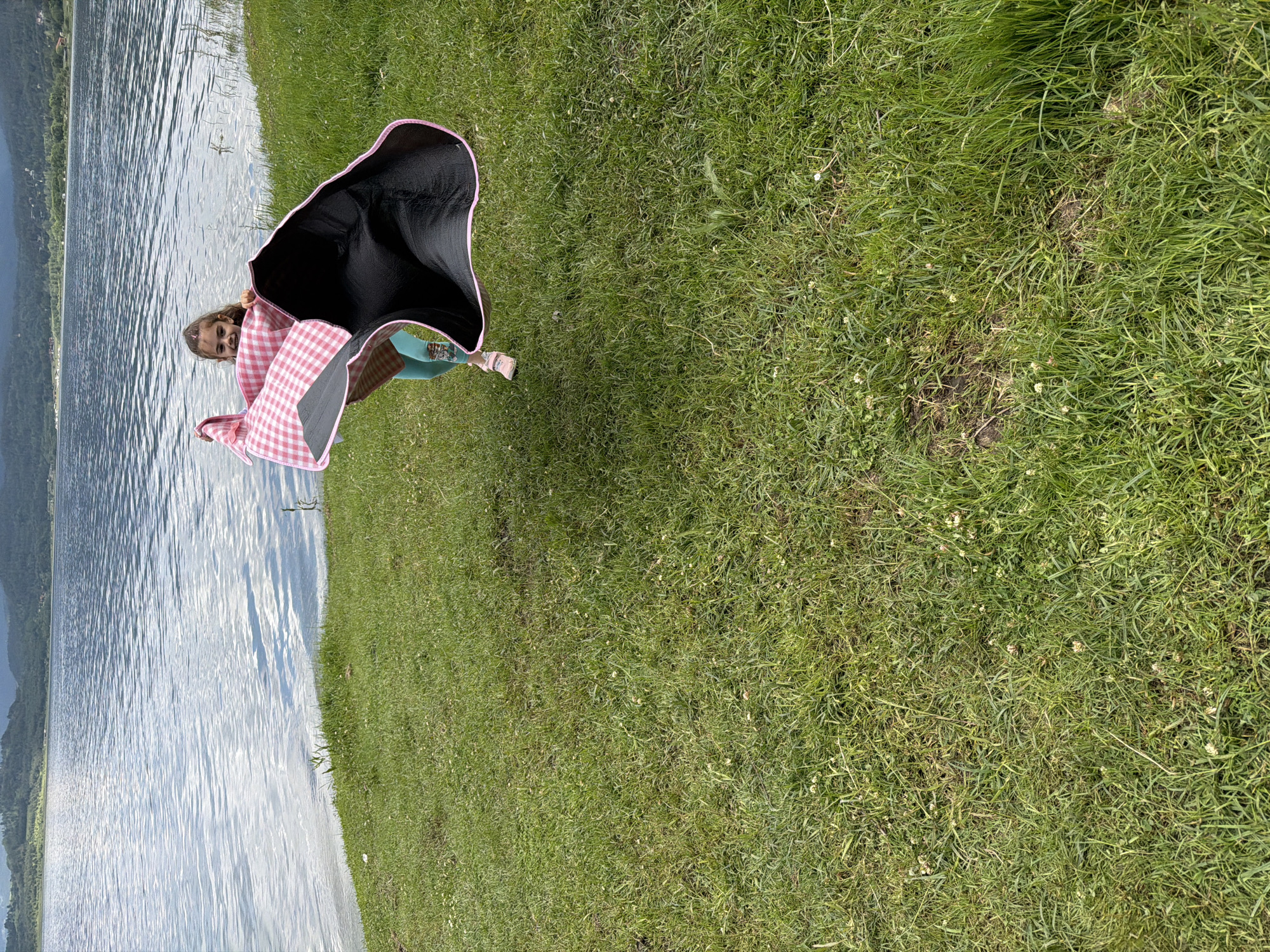 The image captures a full-length outdoor scene featuring a young girl on a grassy bank next to a body of water in Цигов чарк, Bulgaria. The girl, positioned slightly to the right of the frame's vertical center, is holding a large picnic blanket aloft. She has light brown hair tied in a ponytail and is smiling. She wears light blue or teal leggings with a patterned design near the ankles and light pink shoes. The picnic blanket displays a pink and white checkered pattern on one side and a textured black material with pink trim on the reverse side; it is partially unfurled and appears to be catching air.
The foreground and midground consist of a natural, uneven grassy area with varying shades of green, scattered white clover-like flowers, and occasional patches of exposed soil. This grass extends to the edge of a large body of water, likely the Batak Reservoir, which occupies the left side and much of the background of the image. The water surface is rippled and reflects the sky, appearing blue-grey. In the distant background, beyond the water, are tree-covered hills or mountains under what appears to be a bright sky. The overall scene depicts an active moment in a natural, open-air environment.