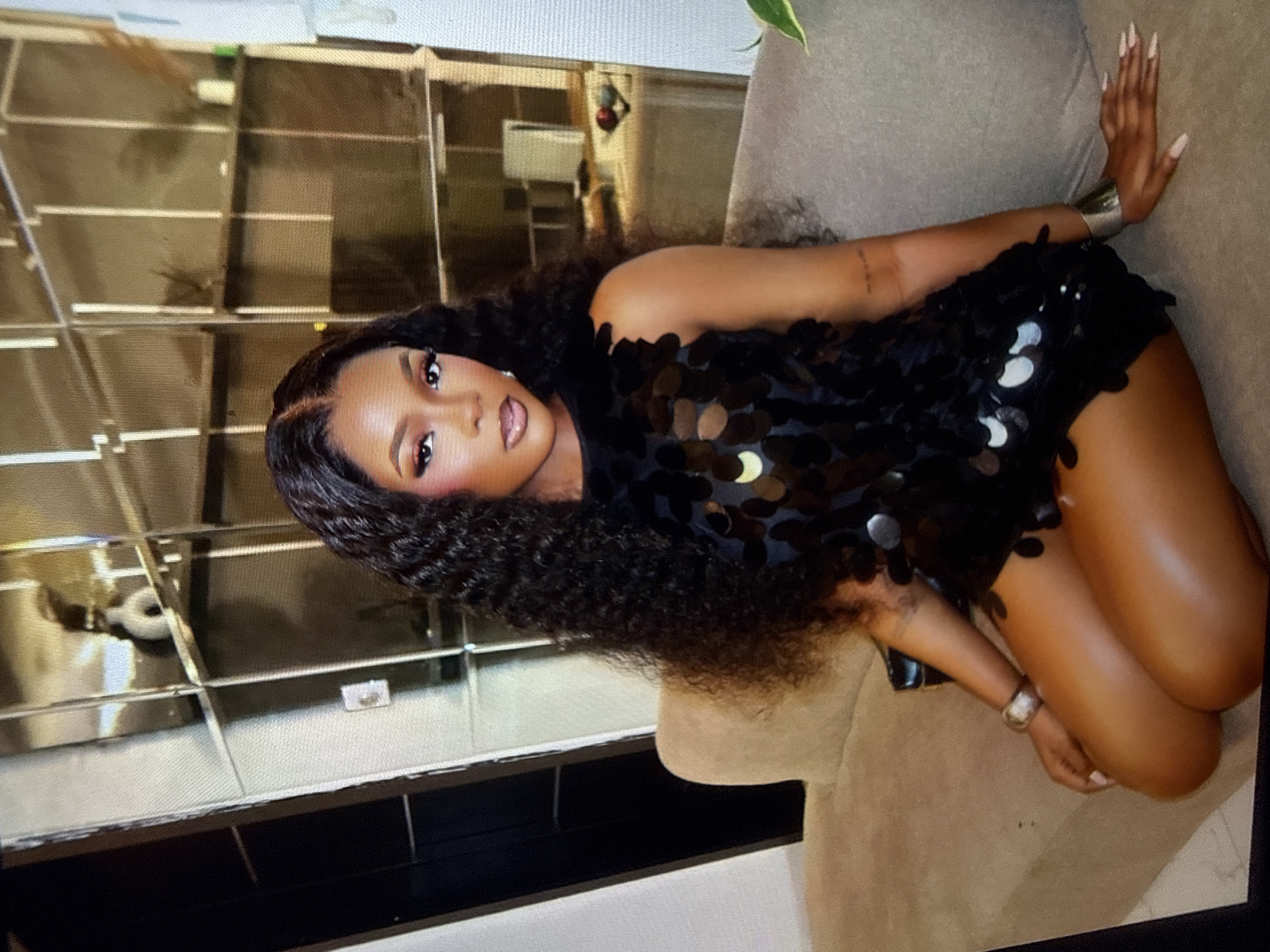 A Black woman with long, dark, curly hair and flawless makeup is sitting on a light brown sofa in what appears to be a modern living room. She is wearing a sleeveless black sequined dress that exposes her legs. She has on silver bracelets on both wrists and her fingernails are painted light pink. The background features a large, floor-to-ceiling shelving unit with multiple compartments, some of which are filled with decorative items. The lighting is soft, casting subtle shadows. The overall impression is one of sophistication and style, suggesting a home in a well-appointed urban setting, possibly in Abuja, Nigeria.