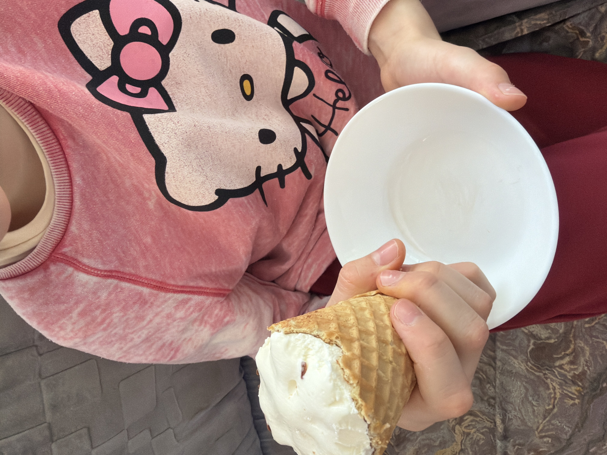 The image shows a person holding an ice cream cone and a white bowl. The person is wearing a pink Hello Kitty sweatshirt and red pants. The background includes a gray cushion and a patterned floor. The location is Кадряково, Russia.