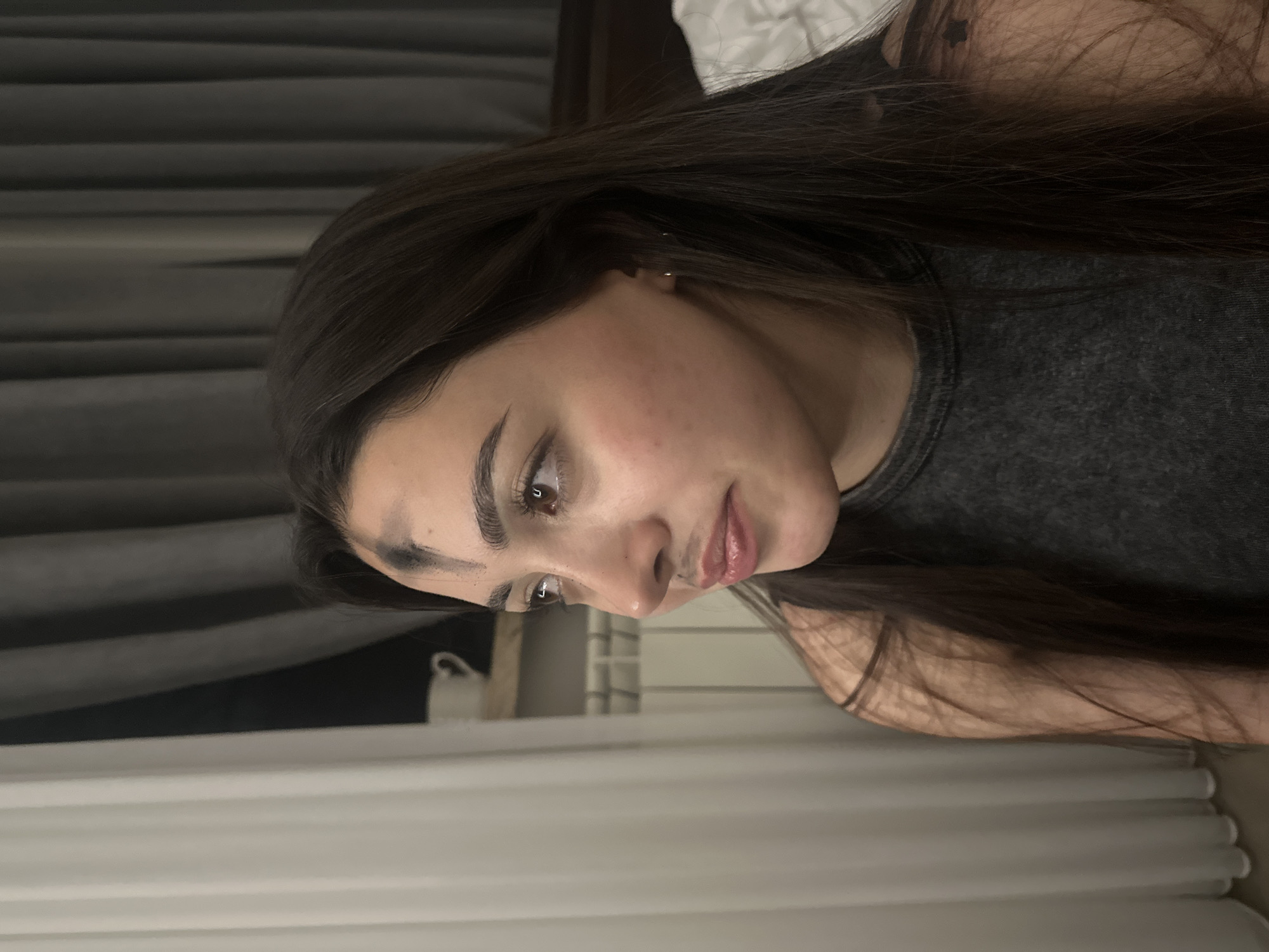 The image depicts a woman with long, dark brown hair, brown eyes, and fair skin. She is wearing a gray tank top. The background includes a horizontally striped dark gray wall and what appears to be a bed with white bedding. A partially visible white object (possibly a lamp or part of a bed frame) is also present. The image does not provide any information about the location being Yerevan, Armenia. The scene is indoors, and the woman is looking downward.