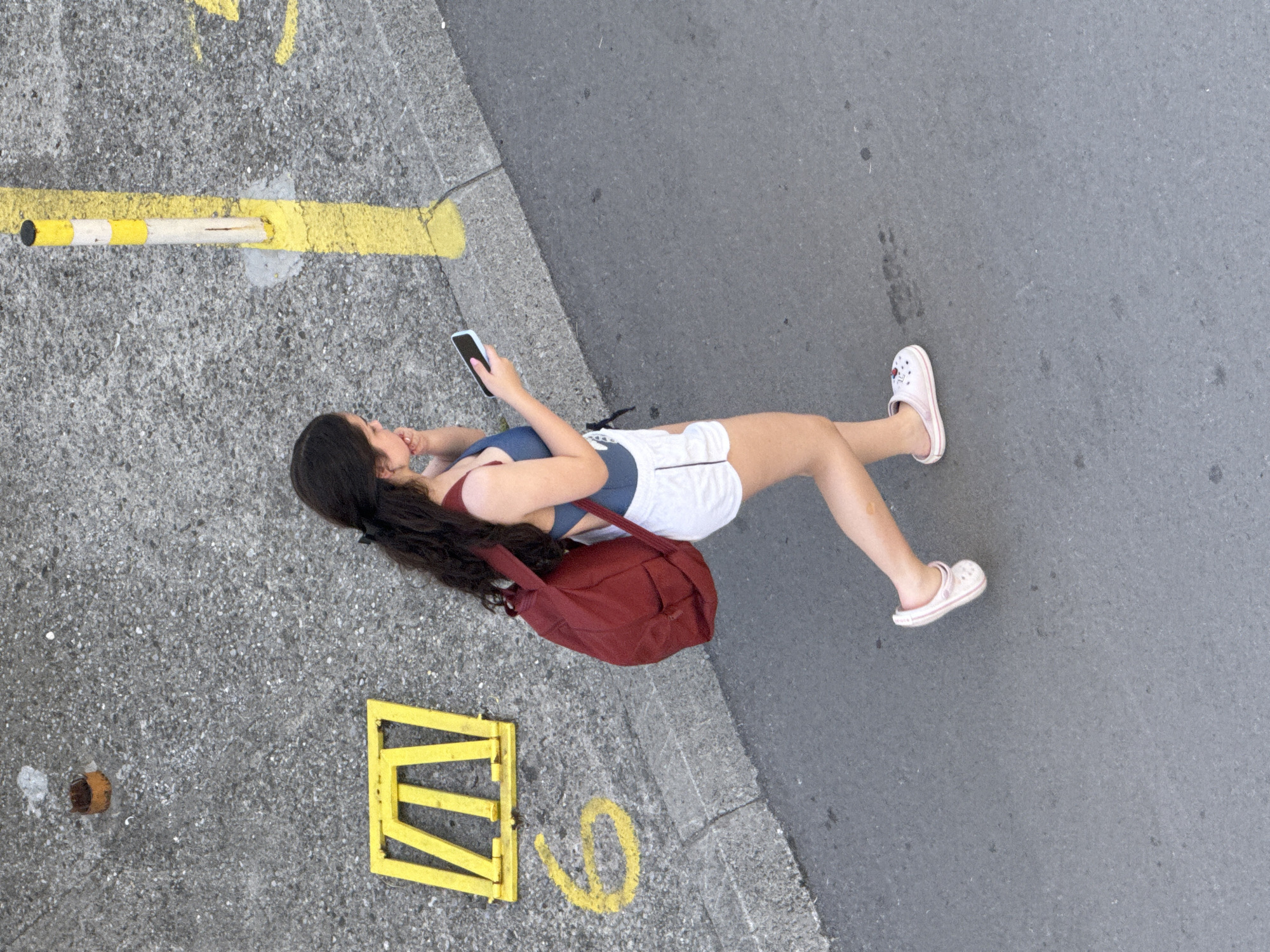 A high-angle shot depicts a young woman on a city street. She is fair-skinned with long, dark hair. She wears a blue athletic top, white shorts, and pink Crocs. She holds a cell phone in her left hand, looking at the screen. A red backpack rests on the ground beside her.
The ground is concrete and asphalt, with faded yellow markings, including a straight line, a geometric shape, and a circular shape. A corroded metal object is embedded in the concrete.
Given the style of clothing, the architecture, and the setting, it is plausible that the scene is located in Budva, Montenegro.