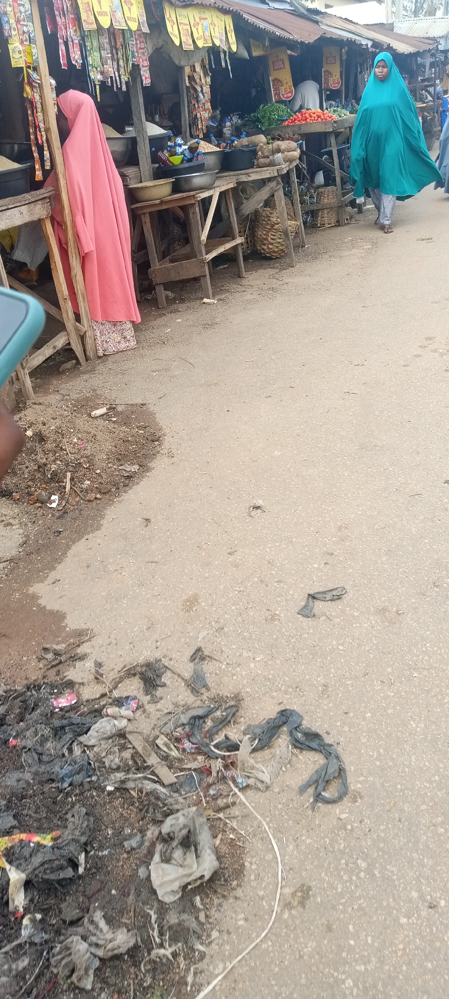 The image depicts a street scene in Jos, Nigeria. Several informal market stalls line the street, selling various goods including vegetables, tomatoes, and possibly rice. Two women are visible, both wearing full-body coverings. One is draped in pink and standing near a stall. Another, in green, is walking down the road. The street is paved but has noticeable amounts of litter including what appears to be discarded plastic bags and other debris. The stalls appear to be constructed of wood and metal sheeting.