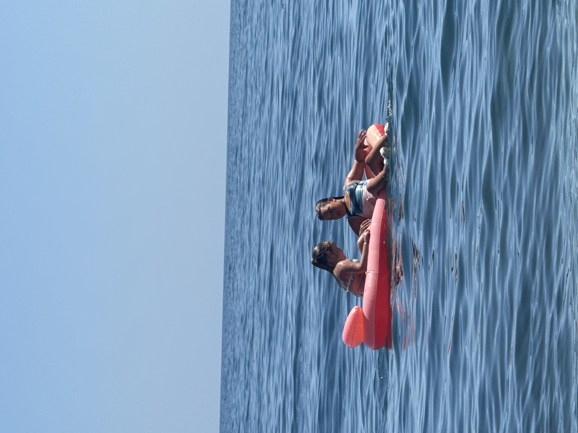 Two young girls are floating on a pink inflatable raft in the sea. The girl on the left has her back to the camera and is looking at the girl on the right. The girl on the right is facing the camera and is smiling. The sea is calm and blue, and the sky is clear and blue. The image appears to have been taken on a sunny day. The location is the city of Utios, Ukraine.