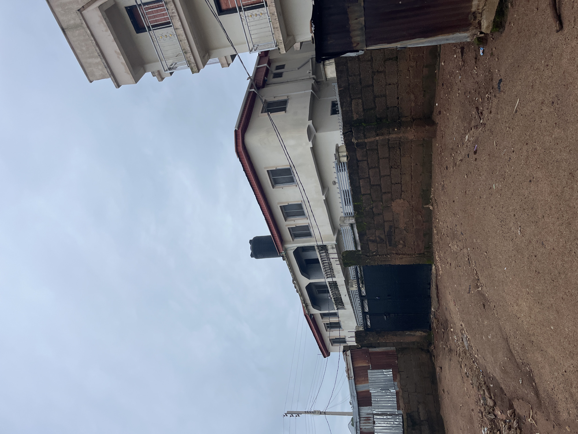 A two-story building with a white facade and red trim is visible, set behind a brick wall and a dark metal gate. The building has multiple windows and balconies, with a large black water tank on the roof. To the right, another part of a building, also white with balconies, is partially visible. In the foreground, a dirt road is present, littered with debris. To the left of the main building, a corrugated metal fence is seen. The sky is overcast. The scene appears to be in a residential area of Jos, Nigeria, characterized by the style of the buildings and the materials used for fencing and construction.