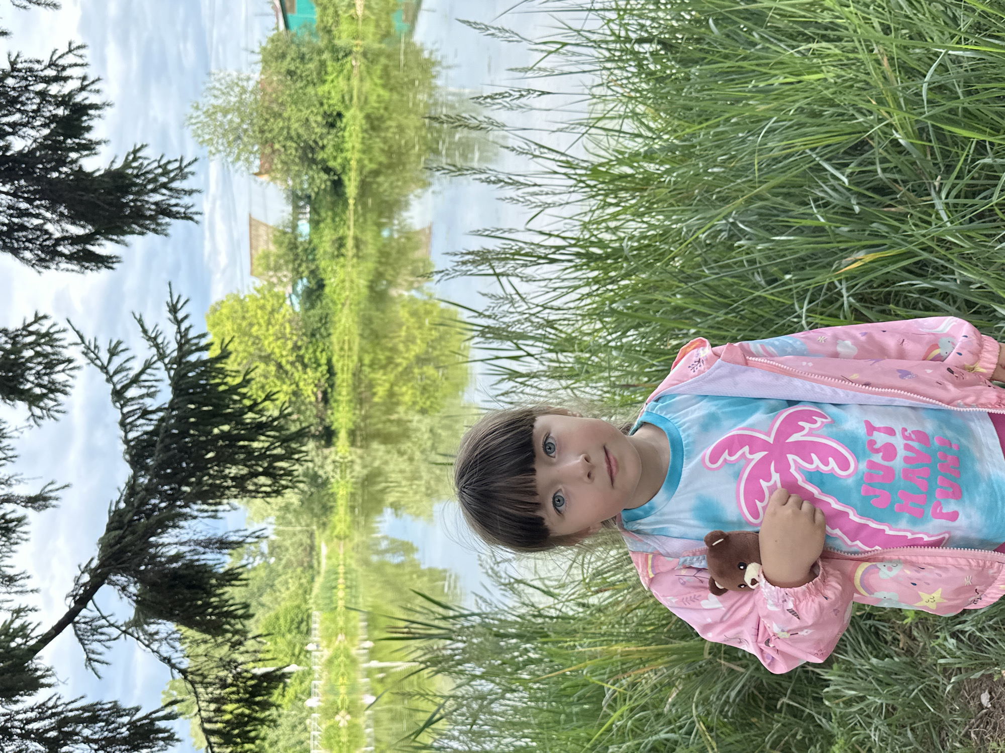 In this vertically oriented photograph, a young girl with blue eyes and dark bangs is the central subject. She is wearing a pink jacket with colorful patterns and a blue tie-dye t-shirt featuring a pink palm tree graphic and the text "JUST HAVE FUN!". She is holding a small teddy bear in her right arm. The girl is positioned in a grassy area, with tall green grass surrounding her. Behind her, a body of water, likely a lake or river, reflects the sky and surrounding greenery, creating a symmetrical scene. The water's surface is calm. Trees, some appearing to be evergreen and others deciduous, line the opposite bank of the water. In the distance, a glimpse of a building with a green roof and a brown roof can be seen amongst the trees. The sky is partially cloudy. The overall impression is a peaceful, outdoor scene, possibly taken during the daytime in a rural or semi-rural setting in Chaikovsky, Russia. The girl's expression is neutral, and the setting suggests a casual, relaxed moment.