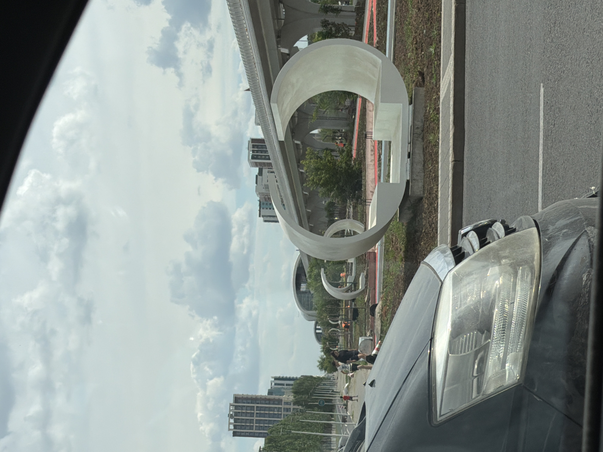 This media file captures a street view from inside a vehicle, looking out towards a modern urban landscape. The scene features a prominent, large-scale abstract sculpture made of white material, resembling intertwined loops or ribbons, situated alongside a road. Behind the sculpture, an elevated structure, possibly a highway or monorail line, is visible with its supporting pillars and track.
In the background, several multi-story buildings with contemporary architecture can be seen, suggesting a developed urban area. Greenery in the form of trees lines the street, interspersed with the architectural elements. The sky is filled with a mix of clouds and clear patches, indicating daytime with potentially variable weather.
Several individuals are present near the sculpture, some appearing to be engaged in construction or maintenance work, with orange cones and equipment visible. Others are walking or standing, observing the surroundings. A black car, from which the photo is taken, is partially visible in the foreground, with its headlight and hood in clear view.
The overall impression is of a city in development or undergoing enhancement, with artistic installations complementing the infrastructure and buildings. The time of day appears to be afternoon or late morning, given the lighting conditions. The location context of Astana, Kazakhstan, suggests a modern Central Asian city. There is no visible text in the image.