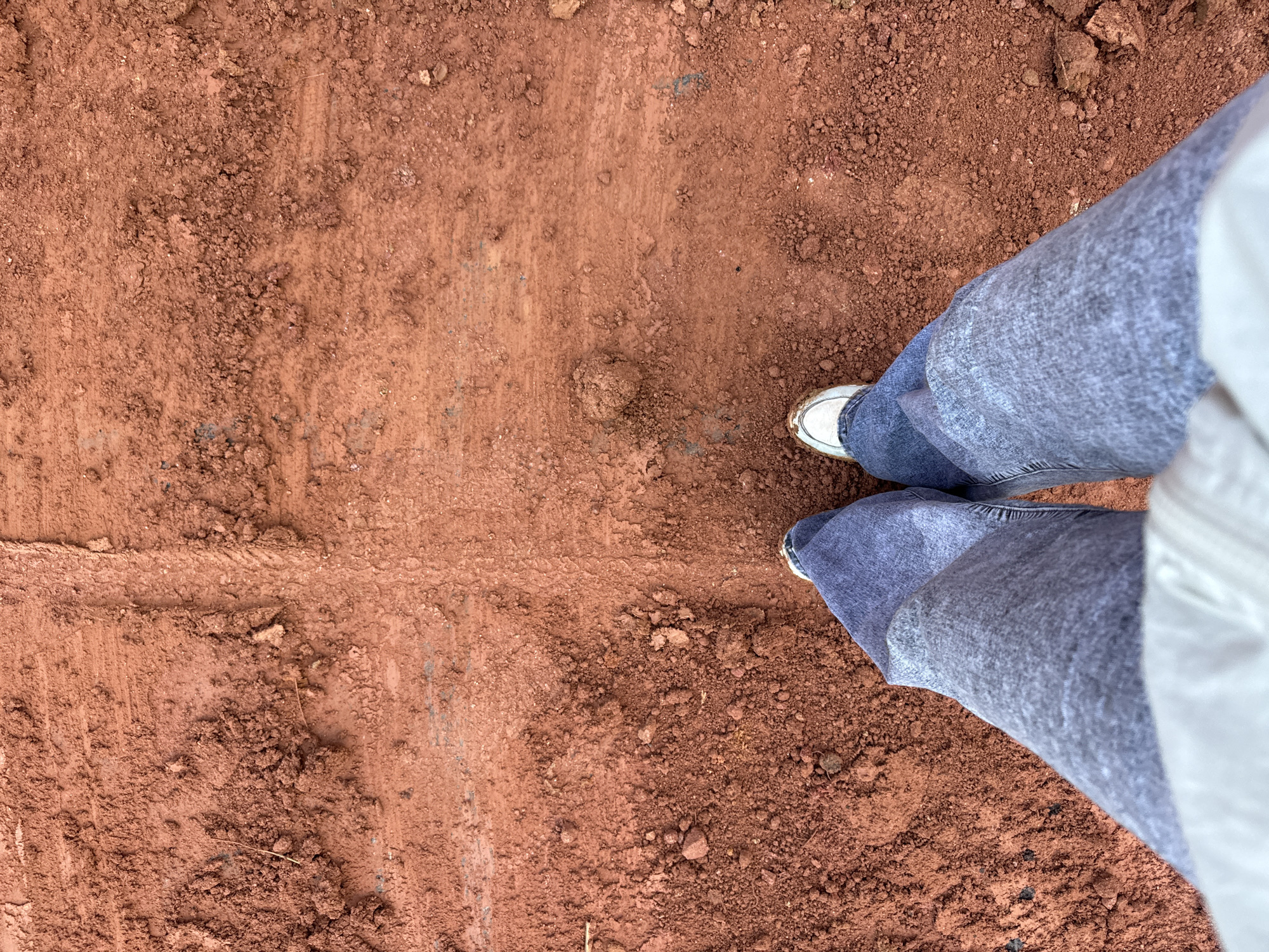The image shows a person's legs and feet standing on reddish-brown dirt, possibly from a construction site or unpaved road in Nao Sẻ, Vietnam. The ground is uneven and appears to have tire tracks imprinted in it. The person is wearing blue jeans and white sneakers. The top of the image shows more of the dirt ground, with some debris scattered around.