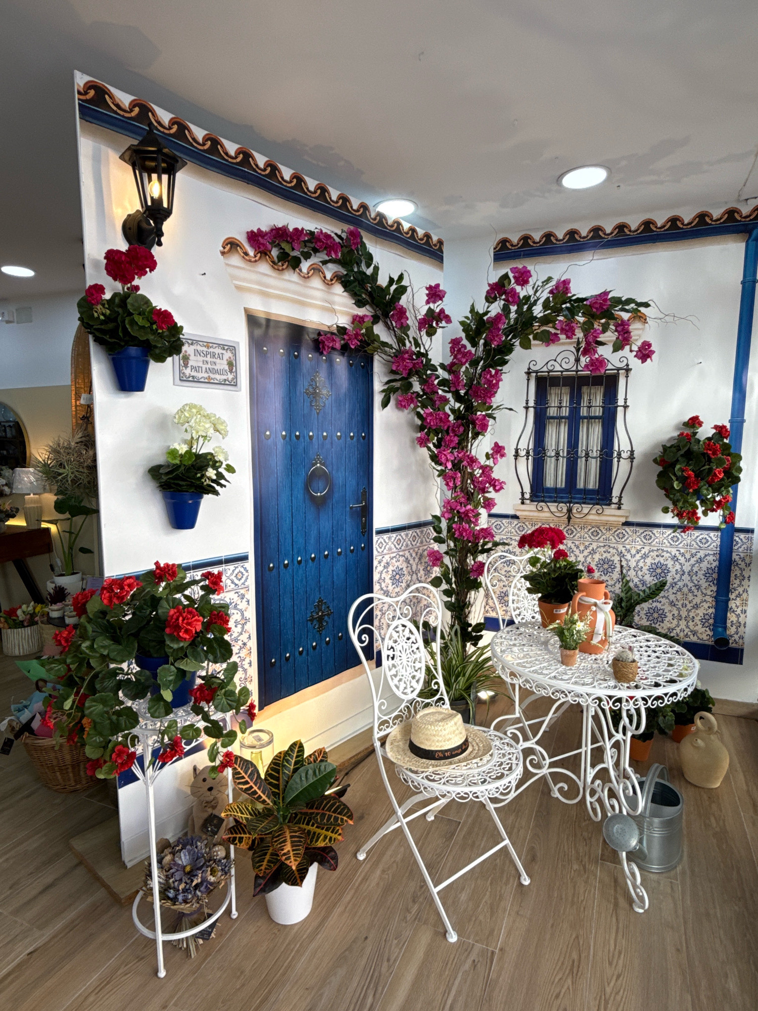 This image depicts a decorative outdoor scene, likely a shop display, in Malgrat de Mar, Spain, designed to evoke a Mediterranean courtyard. A vibrant blue door with decorative studs and a ring knocker is central, flanked by a white wall adorned with blue and white tiled patterns.
To the left of the door, a black outdoor lamp hangs above a blue pot containing bright red flowers. A sign with Spanish text, "INSPIRAT EN UN PATI ANDALUS," is also mounted on the wall. Another blue pot with white flowers sits below the lamp. At the base of the wall, a large pot with red flowers is present, along with a wicker basket filled with colorful items.
To the right of the blue door, a decorative wrought-iron window with blue shutters is visible, framed by a climbing plant with bright pink flowers that extends across the top of the wall. A cluster of red flowers hangs to the right of the window.
In the foreground, a white wrought-iron table and two chairs are set up. A straw hat rests on one chair, and the table displays a terracotta jug, small potted plants, and a cactus. A metal watering can and a light-colored ceramic jug are placed on the floor near the table. The scene is set against a wood-effect floor. The overall impression is one of a charming and colorful Spanish patio.