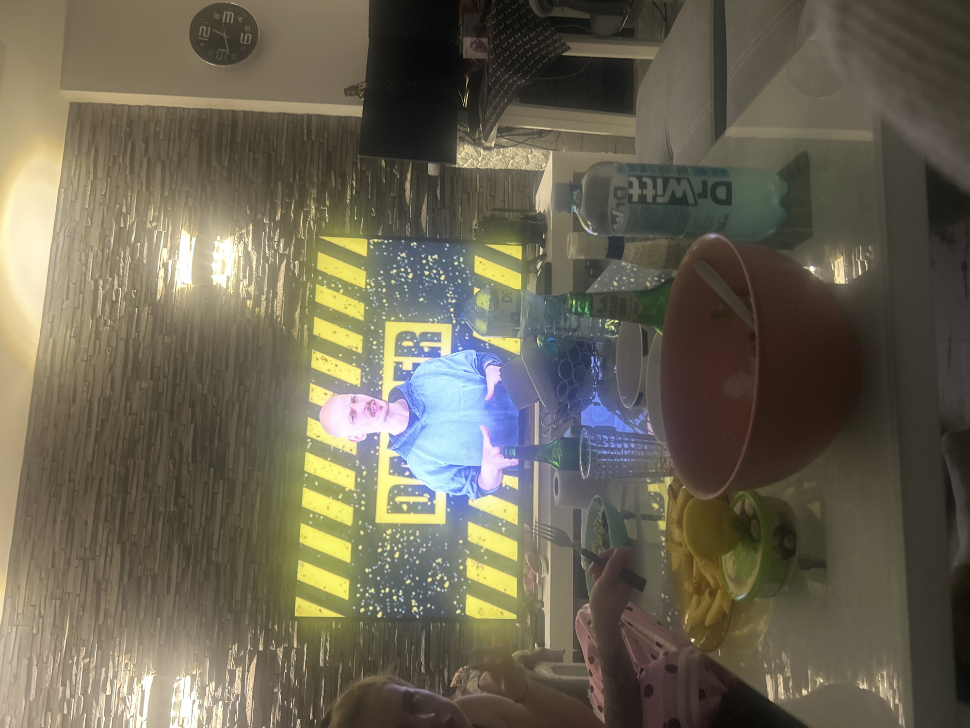 The image appears to be taken inside a home, possibly in Burgas, Bulgaria. A television screen shows a man with a bald head in front of a yellow and black striped background. There is a clock on the wall above what appears to be a decorative stone wall. In the foreground, there is a white table with various objects including a large red bowl, a green Dr. Witt bottle, an assortment of glasses, and a plate with what looks like french fries. There is a person holding a fork, dressed in a pink and black patterned top. The room appears to be dimly lit.
