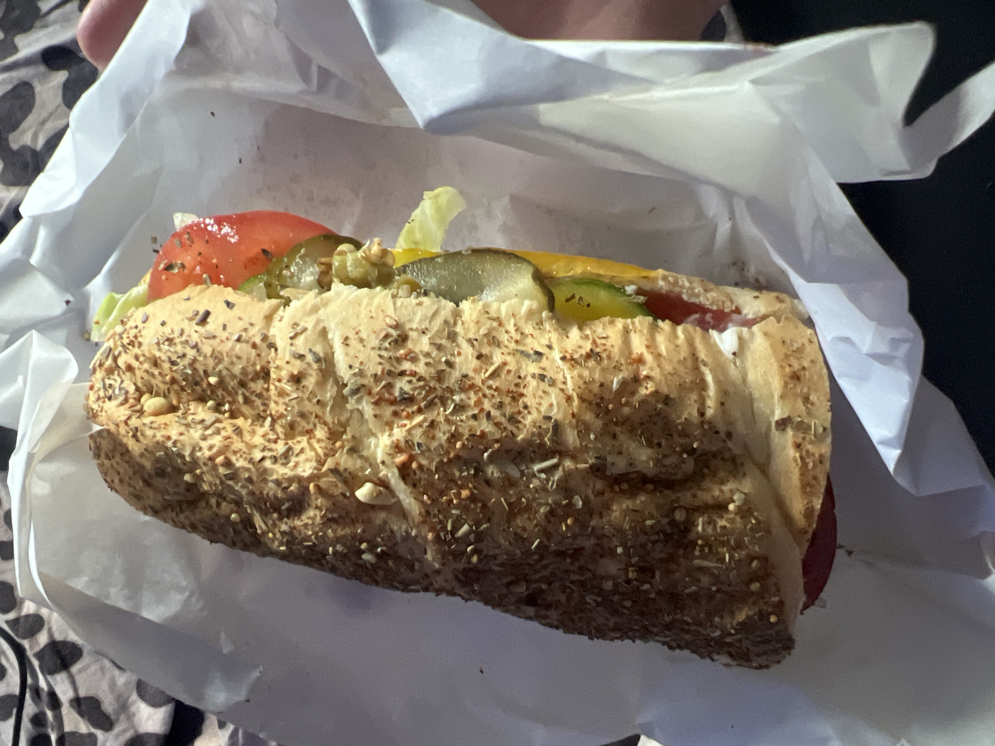 The image displays a close-up view of a sub sandwich, partially unwrapped from its white paper packaging. The sandwich features a textured, multi-grain or seeded bread roll, visibly covered with various seeds and dried herbs on its crust.
Inside the sandwich, several distinct fillings are visible. Slices of vibrant red tomato and fresh green lettuce are prominent, accompanied by green pickle slices. A layer of yellow cheese is discernable, and a creamy white sauce or dressing can be seen. A reddish-brown meat, possibly salami or pepperoni, is peeking out from the lower portion of the sandwich.
The white paper wrapping is open and slightly crinkled, suggesting the sandwich has just been unwrapped or is about to be eaten. The background is out of focus, revealing a blurry pattern of dark and light tones, which could be a blanket or fabric, indicating a casual indoor setting. The lighting appears to be artificial. There is no visible text in the image, and no specific time of day or emotions are discernible. The image is set in Saint Petersburg, Russia.