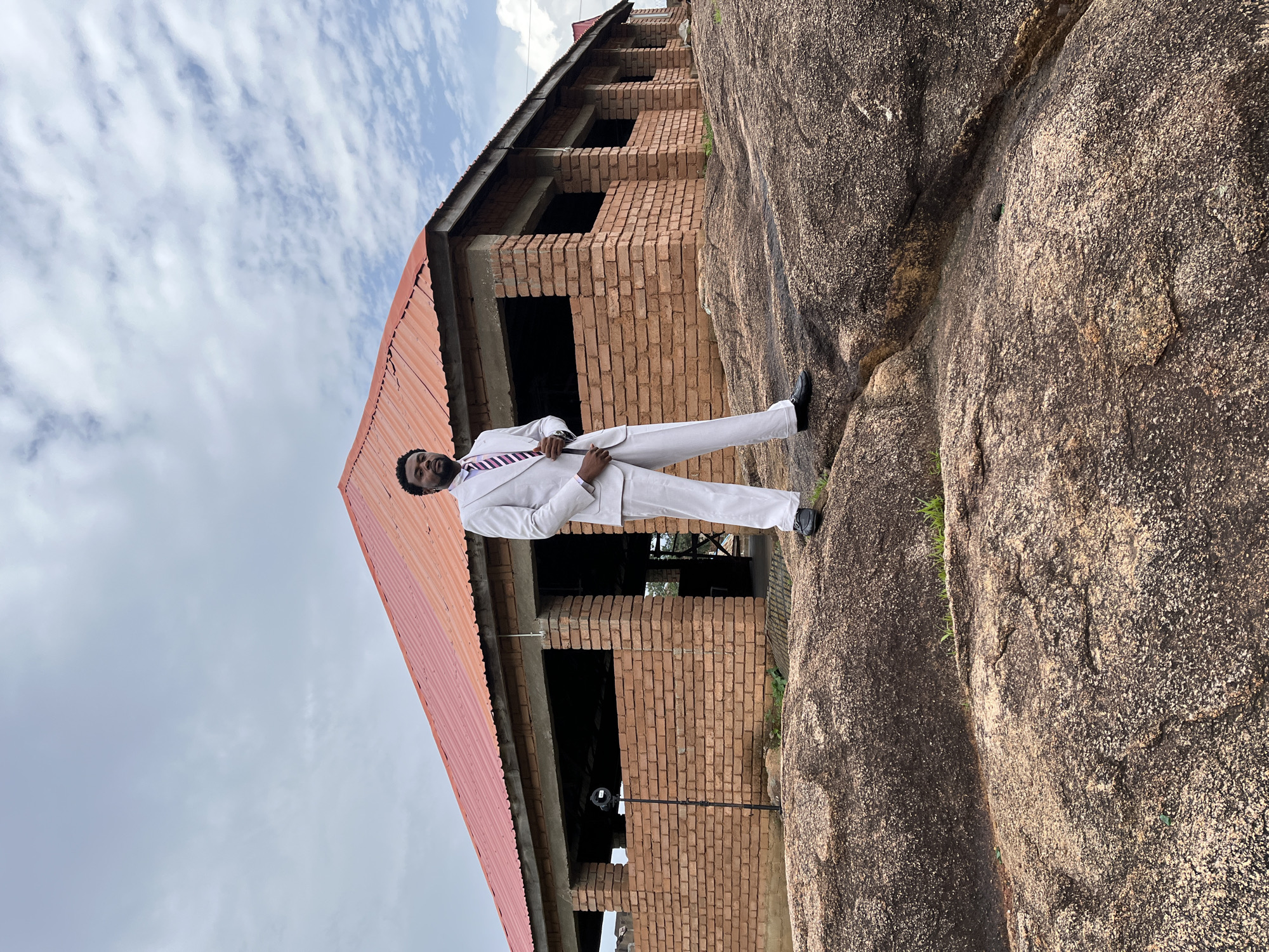 A man in a white suit and a purple and white striped tie stands on a large rock formation in front of a brick building under construction. The building has a red corrugated metal roof and several open window frames. The man is looking towards the camera with a slight smile. The image appears to be taken outdoors during the daytime. The sky is cloudy, and patches of green grass are visible growing from the rock formation. The overall scene suggests a location in Jos, Nigeria, possibly a rural or developing area, given the unfinished building and natural rock landscape. The man's attire suggests a formal occasion or a professional role, despite the rugged surroundings.