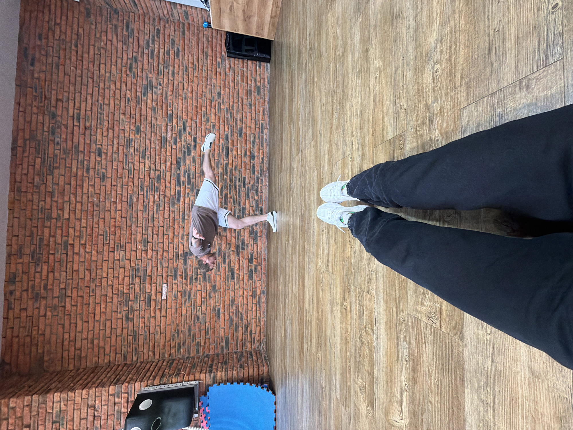 A photo shows the interior of a room, likely a dance studio or fitness area. The image is oriented to make it appear as though the photographer is standing over a person doing a split. The studio is located near a brick wall, potentially in the city of Chaikovsky, Russia, given the urban architectural style typical of Russia.
The room is characterized by a wooden floor. A male figure, dressed in a brown shirt and white athletic shorts, is performing a split along the length of the wall.
In the foreground, two legs covered in black pants with two white sneakers belonging to the person taking the picture are present.
Additional elements within the scene include a section of blue interlocking foam mats, possibly used for cushioning or as a designated exercise area, and a dark rectangular object attached to the wall, likely a TV or a mirror. The setting is indoors, illuminated by unseen light sources.