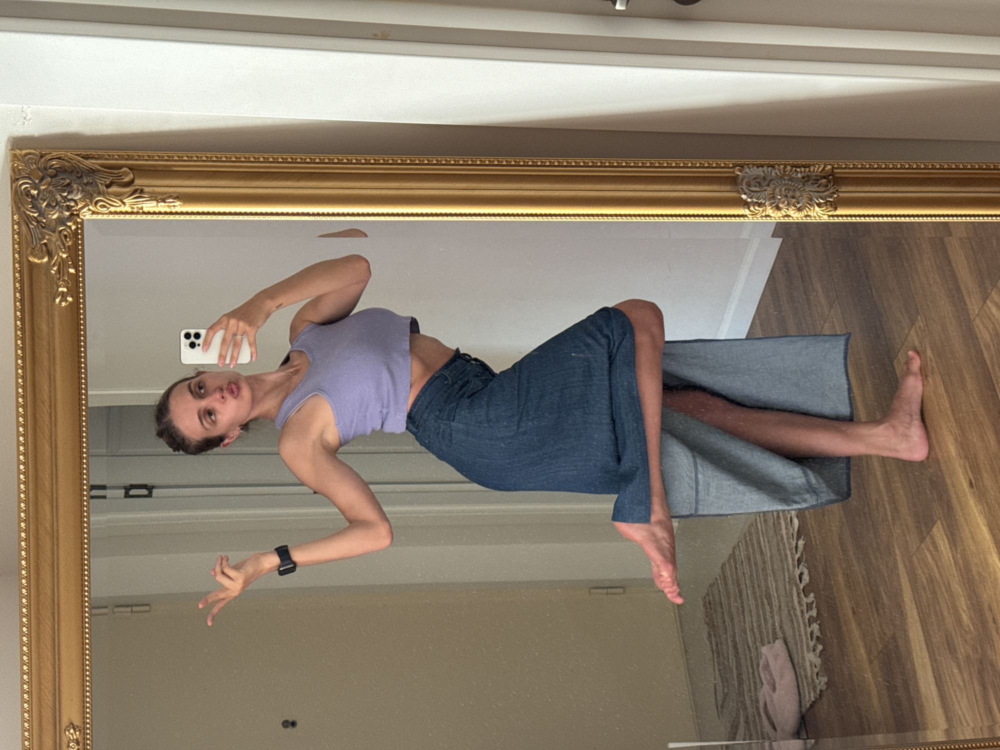 The image, rotated 90 degrees clockwise, displays a mirror selfie of a young woman taken indoors. She stands barefoot in front of a large mirror that features an ornate, golden frame with intricate carvings.
The woman has light skin and her hair is pulled back. She is dressed in a light purple sleeveless crop top, which reveals her midriff, and a dark blue denim garment that appears to be a long skirt or wide-leg pants. She holds a white smartphone in her right hand, using it to capture the reflection. A ring is visible on her right hand's ring finger, and a small tattoo is on her right inner wrist. Her left arm is bent, and her hand is positioned in a distinct gesture. On her left wrist, she wears a black smartwatch. Her facial expression is neutral, with slightly puckered lips, as she looks directly towards the camera.
The mirror's reflection shows the interior of a room. The background consists of light-colored walls and a white ceiling. The lower portion of the reflection reveals a wooden floor with warm-toned planks. A light-colored, possibly braided rug with fringes is partially visible on the floor, along with a soft pink cloth resting on it. The overall setting suggests a casual, domestic environment. No discernible text is visible within the image.