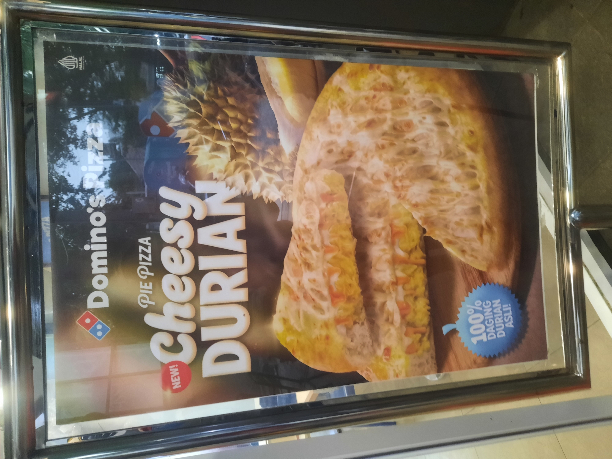 A poster for Domino's Pizza's "Cheesy Durian Pie Pizza" is displayed in a metal frame. The poster features a large image of the pizza, topped with cheese and what appears to be durian. The Domino's logo is visible at the top, along with text that reads "NEW!" and "PIE PIZZA." A durian fruit is also pictured next to the pizza. In the bottom left corner, a blue circular emblem states "100% DAGING DURIAN ASLI!" which translates to "100% REAL DURIAN MEAT!". The poster is framed by a reflective metal border and appears to be indoors, possibly in a restaurant or retail space in North Jakarta, Indonesia, given the presence of the "HALAL" logo, common in Indonesian food establishments. The lighting suggests an interior setting with overhead illumination.