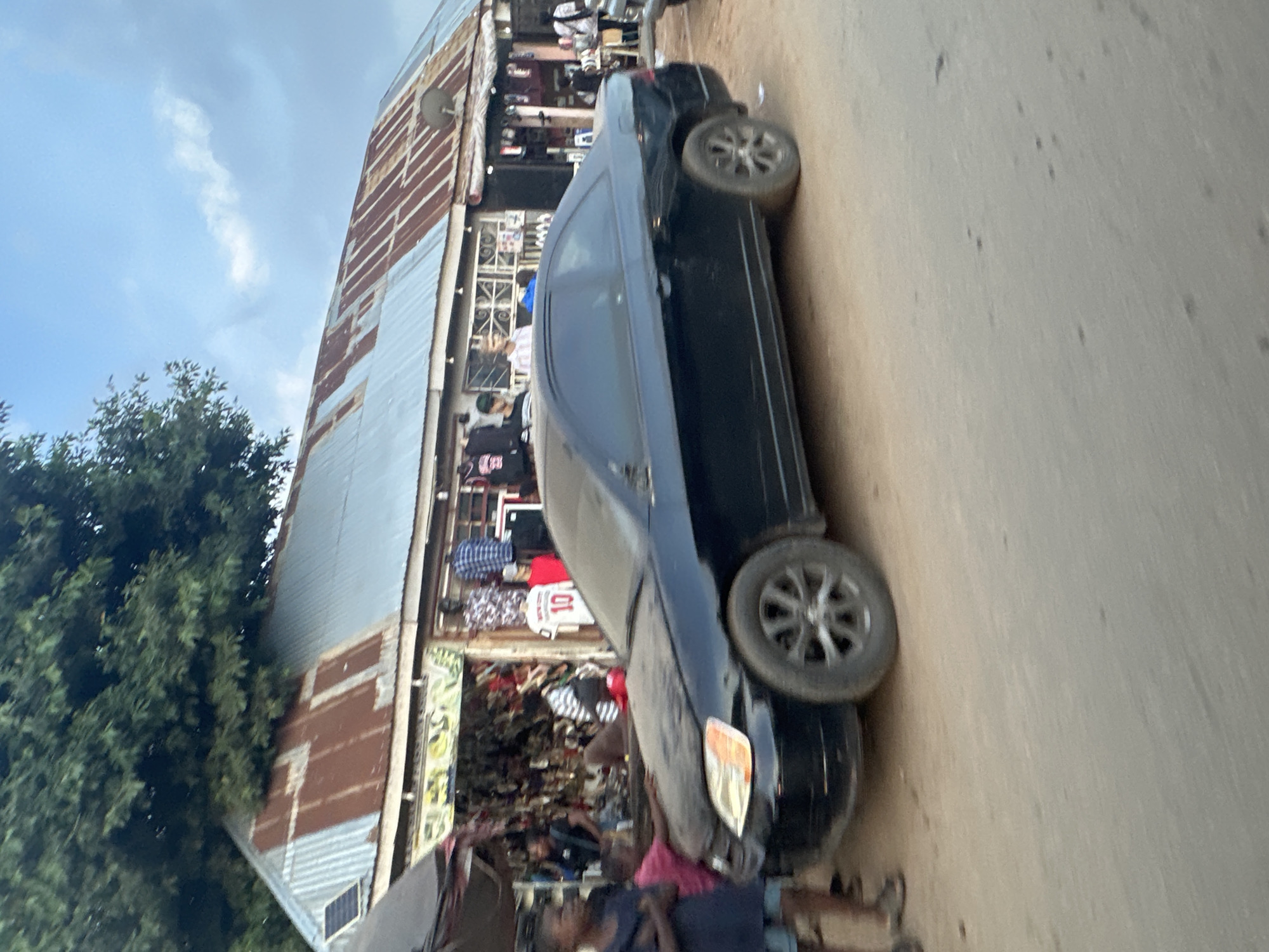 The image shows a street scene in Abuja, Nigeria, with a black coupe parked on a dirt road in front of a row of shops. The shops have corrugated tin roofs, some of which are rusted. Various goods are displayed outside the shops, including clothing and what appear to be shoes or bags. A few people are visible in the image, including children standing near the car and adults browsing or working in the shops. The sky is cloudy. The car's front passenger-side fender appears to be damaged.