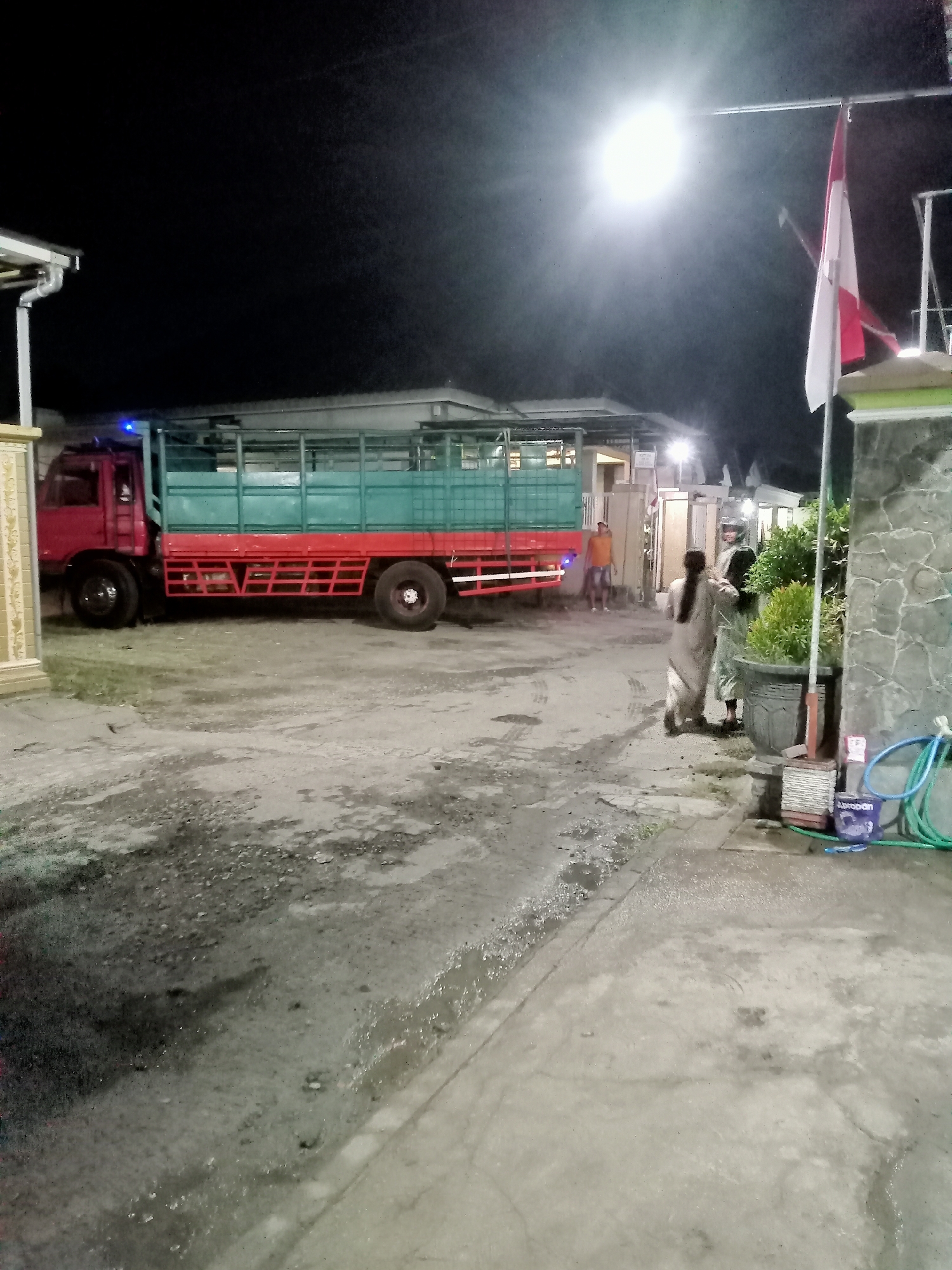 In the city of Simo, Indonesia, at night, a red truck with a green cargo bed is parked on a wet, unpaved area. The truck's bed is filled with what appears to be hay or straw. To the right of the truck, two individuals, one wearing a light-colored head covering and the other in lighter clothing, are interacting near a potted plant. In the background, a building with a tiled roof is visible, and a flag, red and white, is prominently displayed on a pole above the building. A bright street light illuminates the scene. The ground in front of the truck shows tire marks, indicating recent movement.
