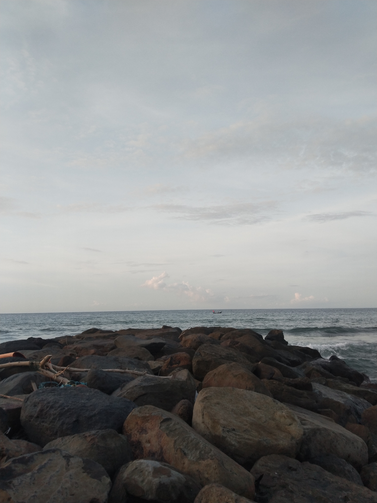 The image depicts a seascape at Lhokseumawe, Indonesia. Large rocks form a breakwater or shoreline barrier in the foreground. The rocks vary in size and color, ranging from dark gray to brown. Beyond the rocks, the ocean extends to the horizon under a cloudy sky. Waves are visible, breaking along the shoreline. A small boat is discernible on the water in the distance. Some pieces of driftwood and debris are scattered among the rocks.