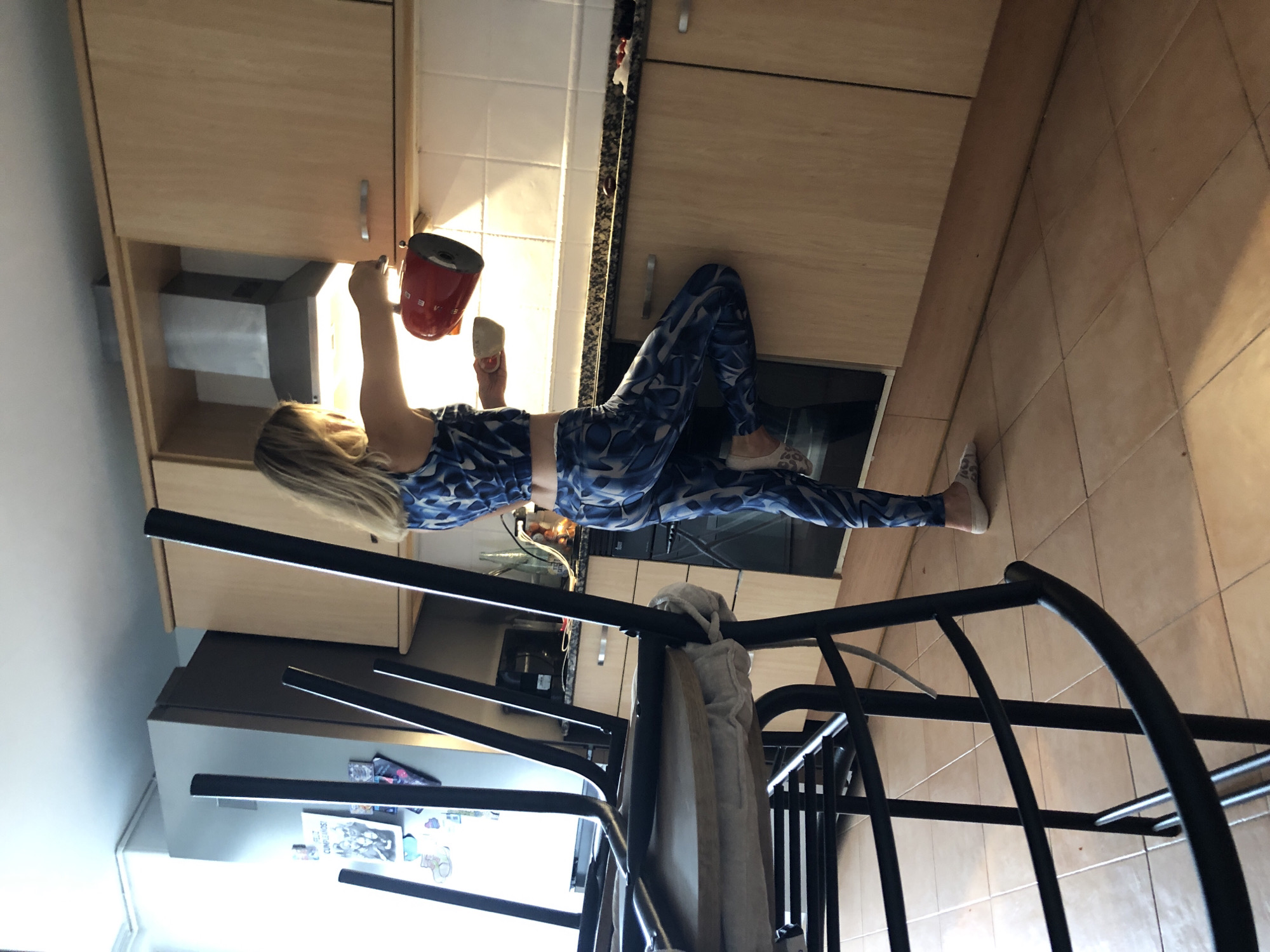 A woman in blue patterned leggings and a matching sports bra stands in a kitchen, holding a red Smeg kettle. She is reaching for a white cup. The kitchen has light wood cabinets and tan tiled floors. A black metal shelving unit is in the foreground, with a beige robe hanging on it. Sunlight streams into the room, suggesting it is daytime. The image is oriented vertically. There is some visible text on a piece of paper attached to the shelves, but it is not legible. The overall scene appears to be casual and domestic, possibly indicating the woman is preparing a drink or cleaning in her kitchen.