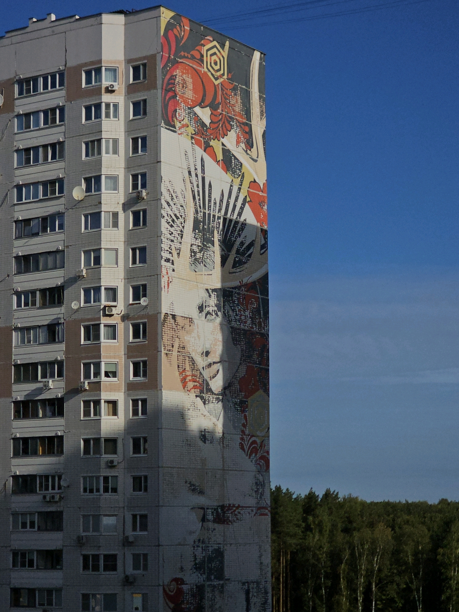 A tall, multi-story apartment building dominates the left side of the frame, its facade adorned with a large, colorful mural. The mural depicts a stylized portrait of a woman with dark hair, her face partially in shadow cast by the setting sun. The building itself is grey with numerous windows and balconies, typical of Soviet-era architecture. A strip of dark green trees forms a treeline along the bottom right of the image, with the bright blue sky filling the remaining space. The building is located in Odintsovo, Russia.