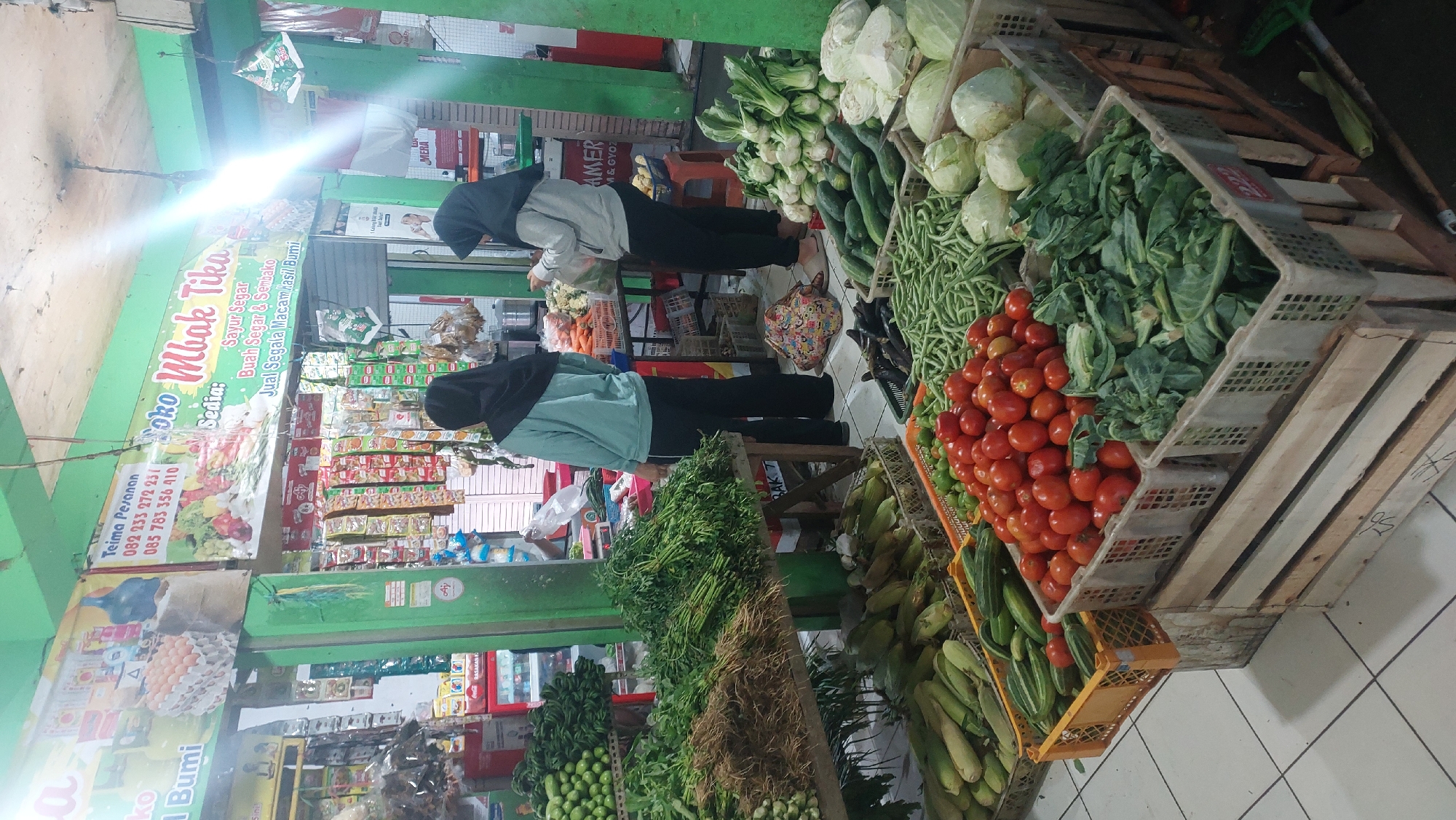 This image shows an indoor market stall in Malang, Indonesia. The stall is filled with a variety of fresh produce, including tomatoes, cucumbers, green beans, cabbage, corn, and leafy greens. Two women, both wearing headscarves, are visible at the stall. One woman is standing behind the counter, and the other is examining some produce. The stall has a sign that reads "Toko Mbak Tika" and lists phone numbers for orders. The environment appears to be a bustling market with other stalls and customers in the background.
