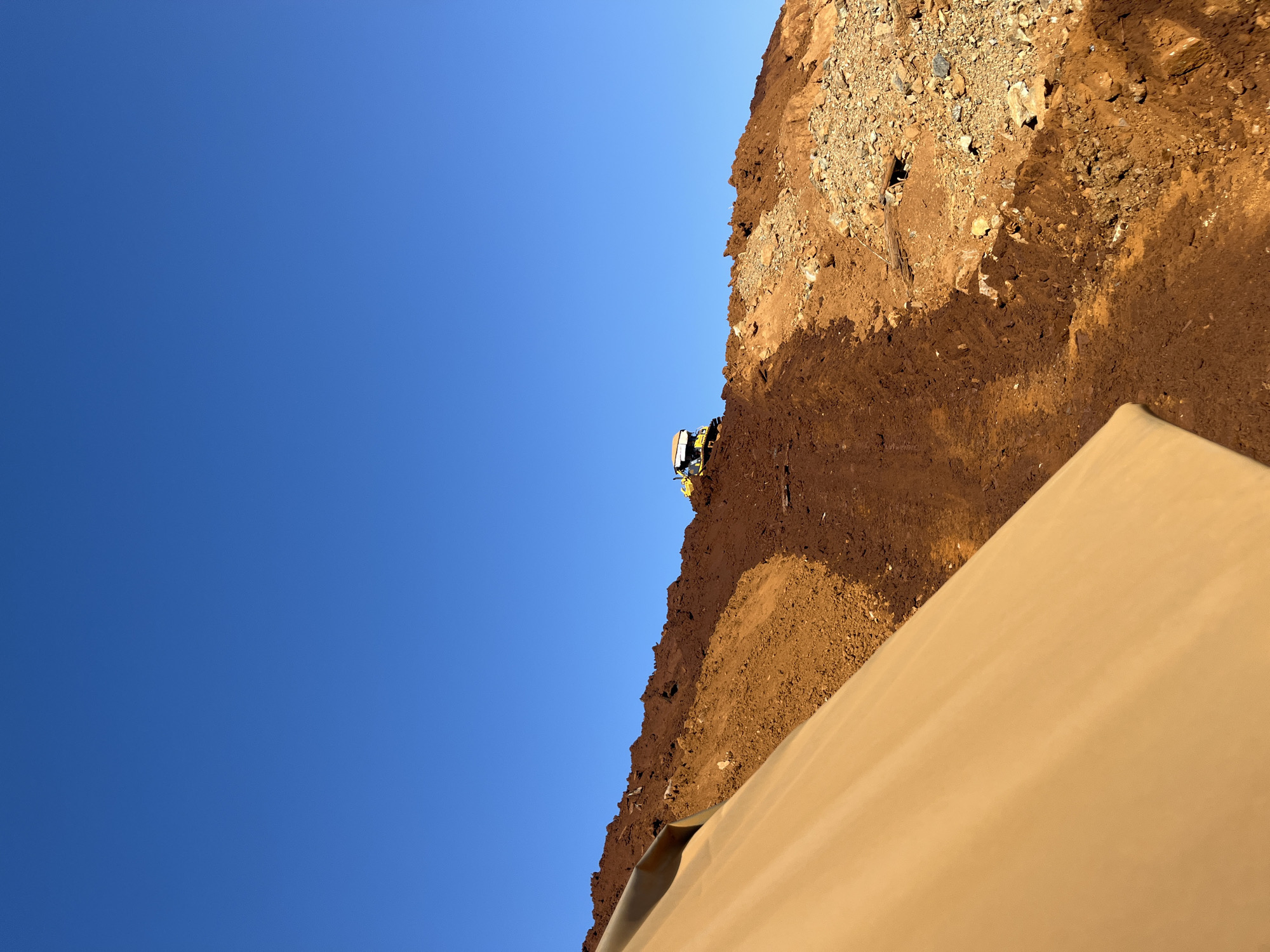 A bright, clear blue sky dominates the upper portion of the image. The lower portion depicts a hilly, excavated landscape covered in various shades of soil and rocks. In the middle ground, a yellow construction vehicle, possibly a bulldozer or excavator, is operating on the slope. In the foreground, a large, tan-colored tarp or fabric is draped diagonally across the frame, partially obscuring the view of the earth. The scene appears to be a construction or excavation site under development, likely in an outdoor, undeveloped area. The specific location in the city of Unknown, Indonesia is not discernible from the visual information.