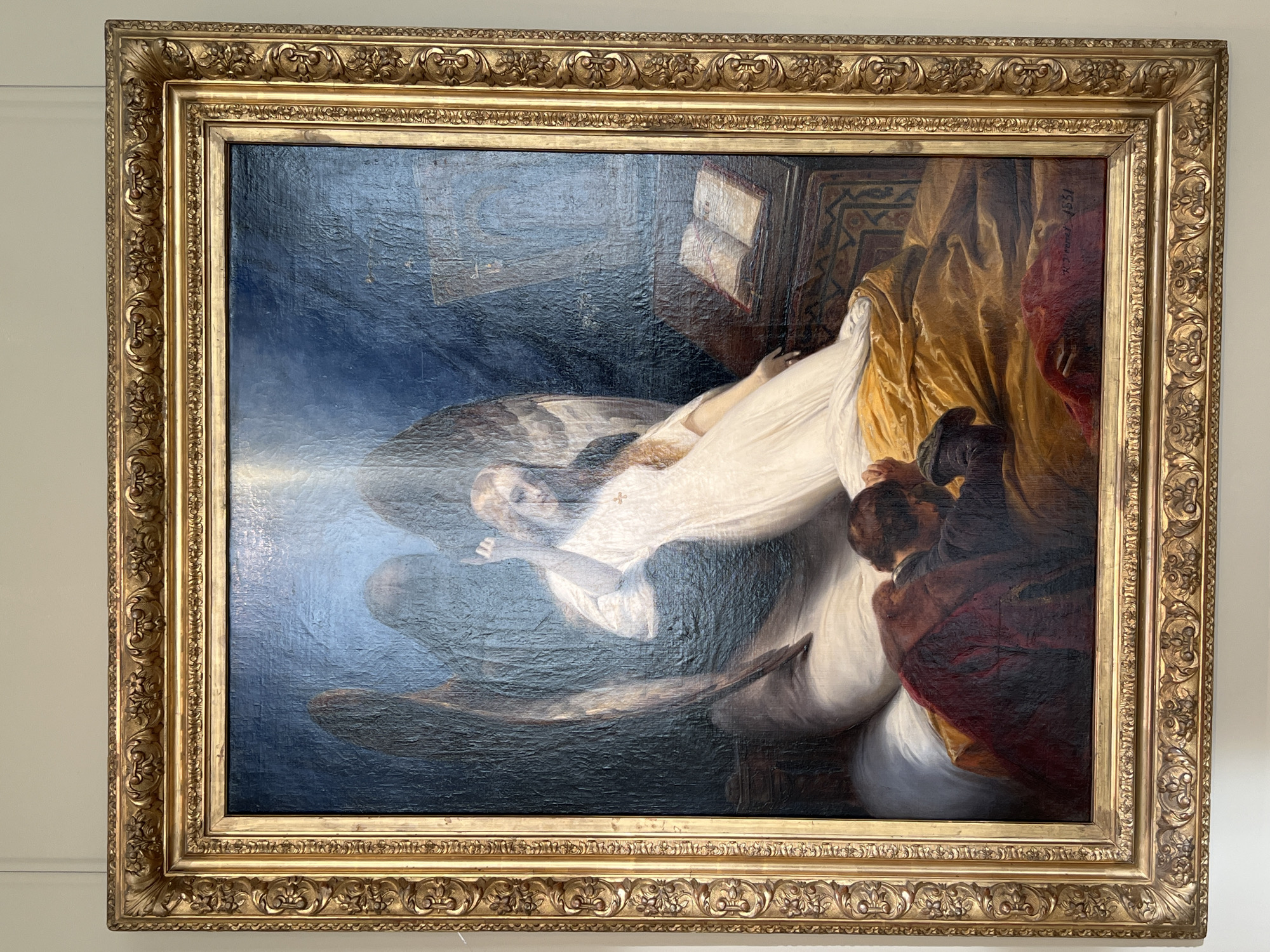 A framed oil painting depicting a woman lying in bed, attended by two figures, against a dark background. The woman is dressed in white and appears to be reclining with an open book and ornate bedding nearby. Two dark-haired figures flank the bed to her right, shrouded in dark robes. The painting is held within a gold-colored frame featuring ornamental details. The inscription "K.P. Briullov 1851" is visible on the right side of the painting. The painting is on display in Saint Petersburg, Russia, likely in a museum.