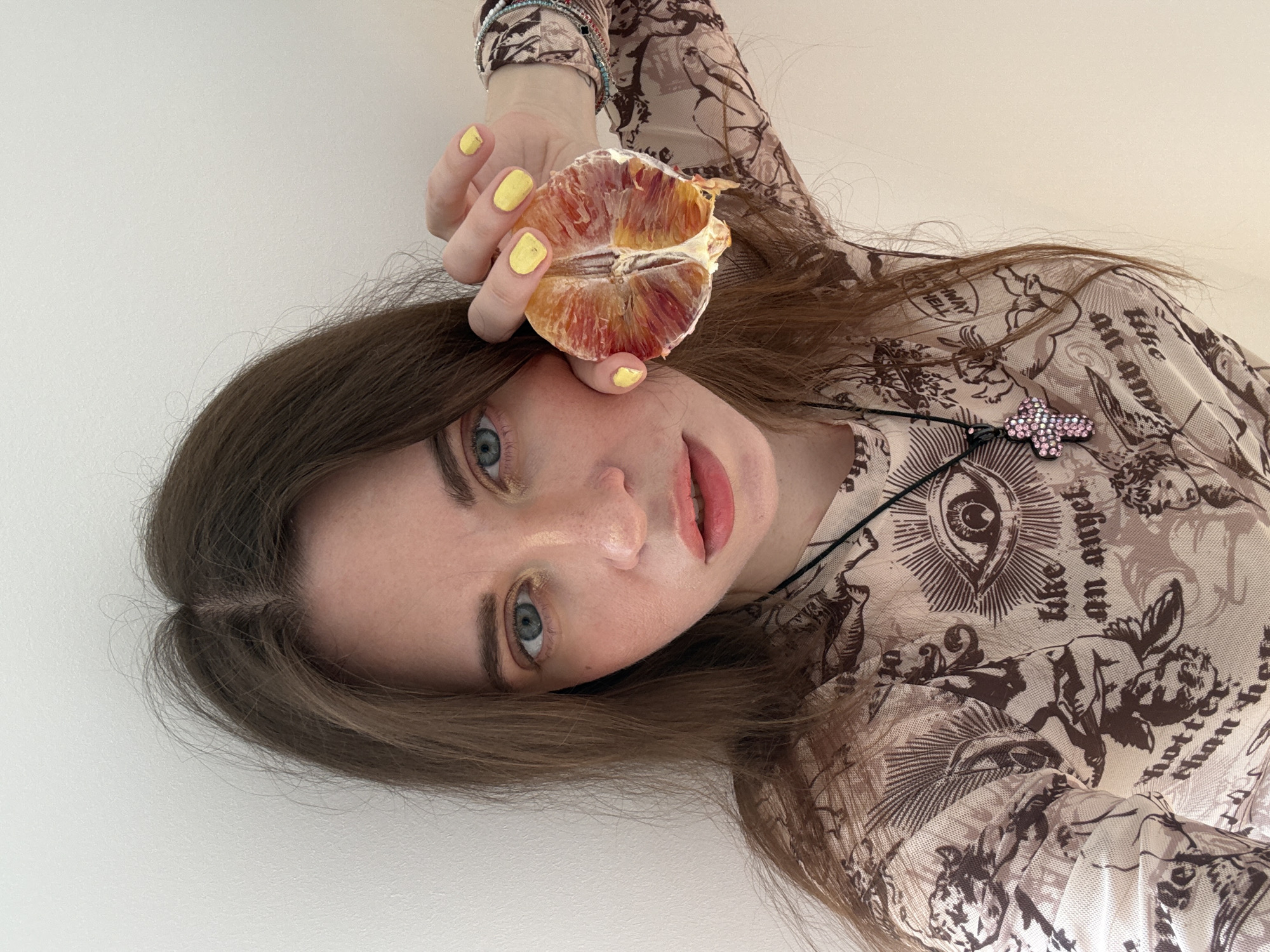 A woman is holding half of a blood orange in her right hand near her face. She has shoulder-length brown hair and blue eyes. Her nails are painted yellow. She is wearing a sheer, long-sleeved shirt with a pattern of angels and eyes. A cross pendant hangs around her neck. The background is a plain white wall. The image is oriented at an angle. There is no indication this photo was taken in Moscow, Russia.