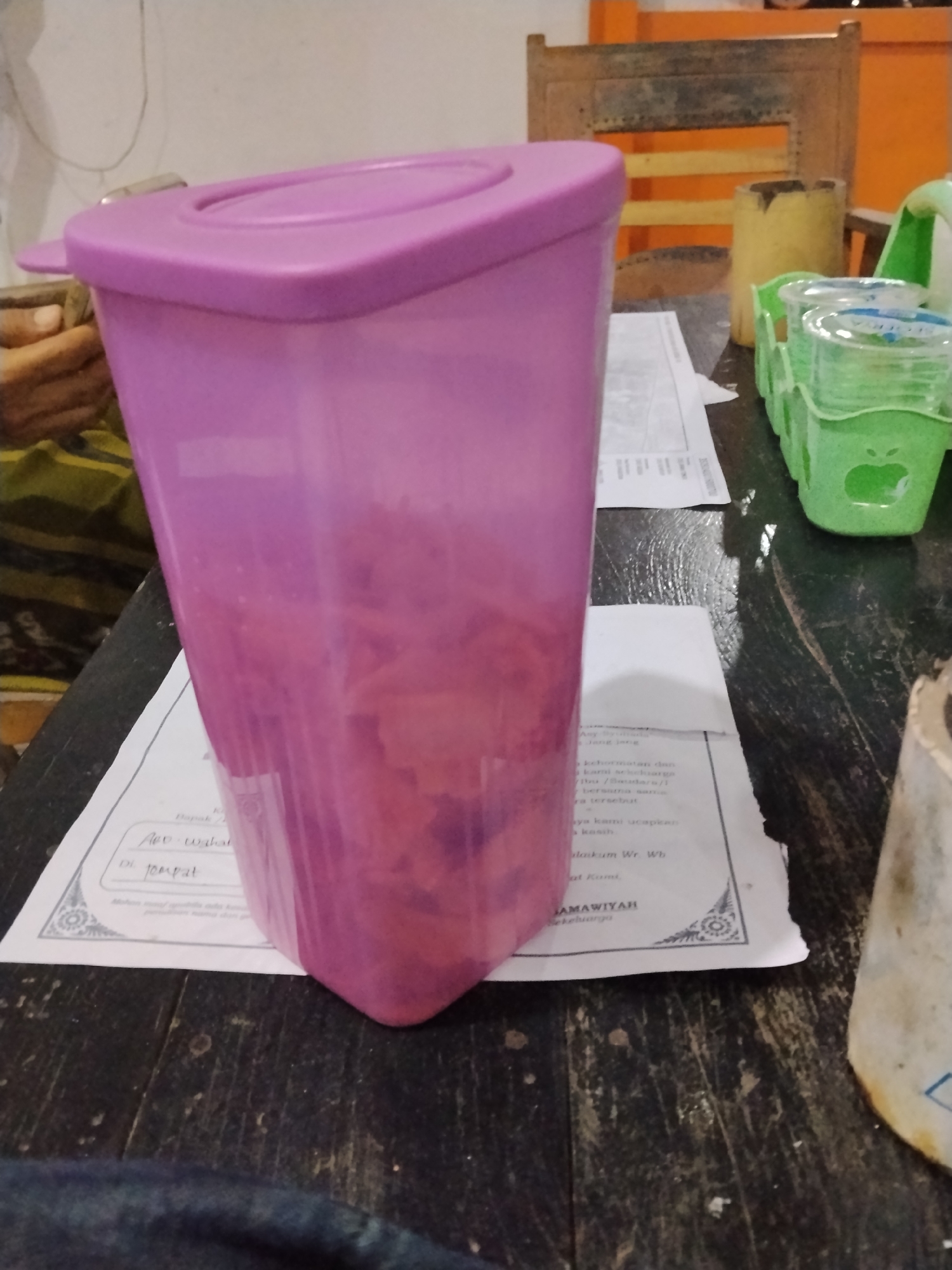 A close-up, low-angle shot captures a tall, translucent pink plastic container filled with a pinkish substance, possibly food, resting on a dark, wooden surface. The container has a purple lid with a handle. To the right of the container, two green plastic holders, each with three small cups inside, are visible. These cups appear to contain small amounts of a clear liquid. In the background, a wooden chair with orange accents and a yellow cylindrical object are present. A white piece of paper with Indonesian text is partially visible under the pink container. The scene is indoors, with a plain white wall in the background. The lighting suggests a dim, artificial environment, possibly at night. The image does not provide any information about the location in the city of Bilis Bilis, Indonesia.