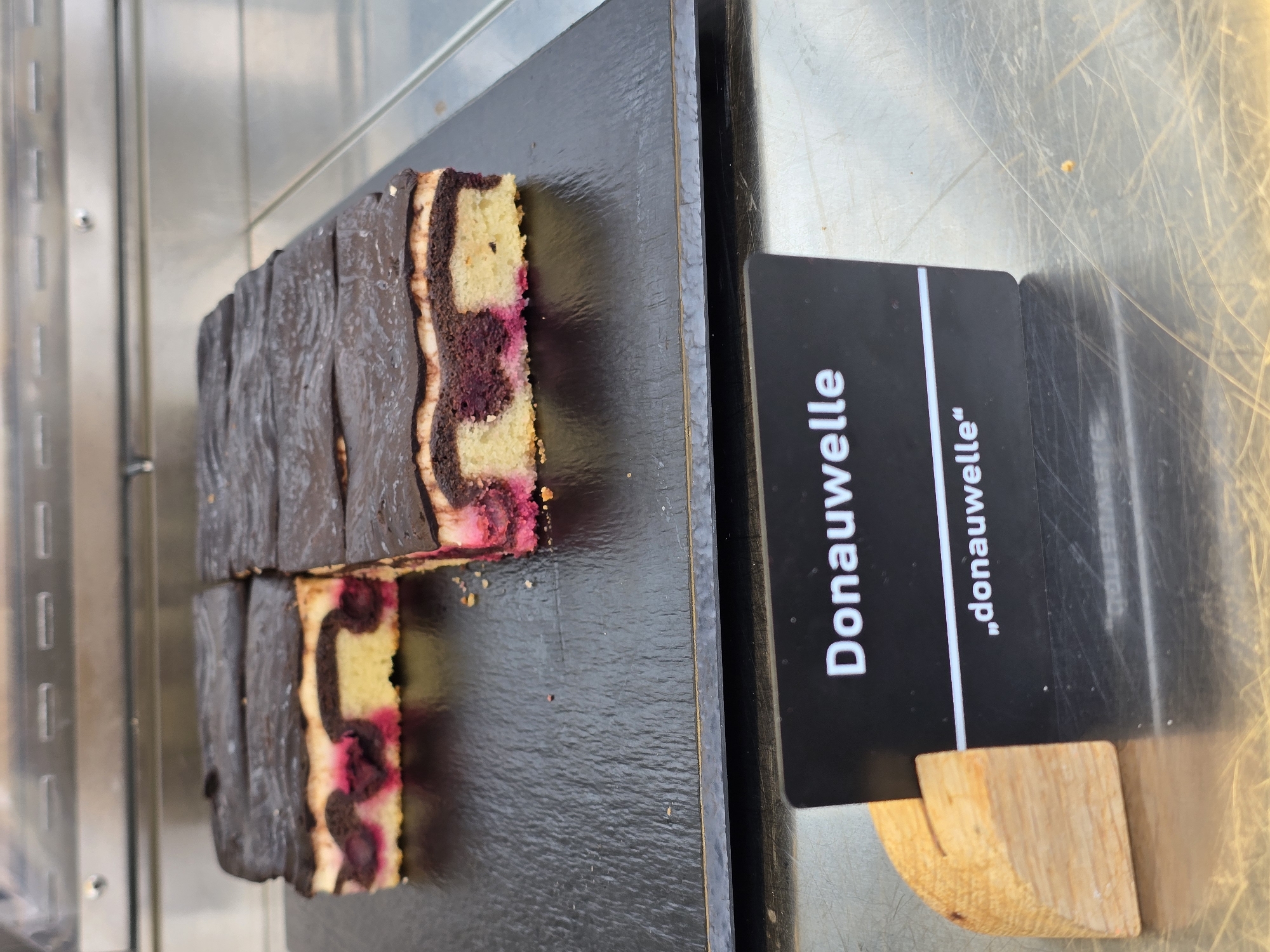 The image shows a close-up view of several slices of a "Donauwelle" cake, a traditional German pastry, displayed on a dark surface, likely in a bakery or cafe in Garmisch-Partenkirchen, Germany. The cake has a marbled batter with dark chocolate and yellow sponge cake layers, interspersed with bright pink cherries or cherry filling. The top is coated with a dark chocolate glaze. A black sign with white lettering, displaying "Donauwelle" and in quotes "donauwelle", is propped up by a small wooden stand in the foreground, obscuring part of the cake. The background appears to be a stainless steel display case. The scene is well-lit, highlighting the textures and colors of the cake and the signage.