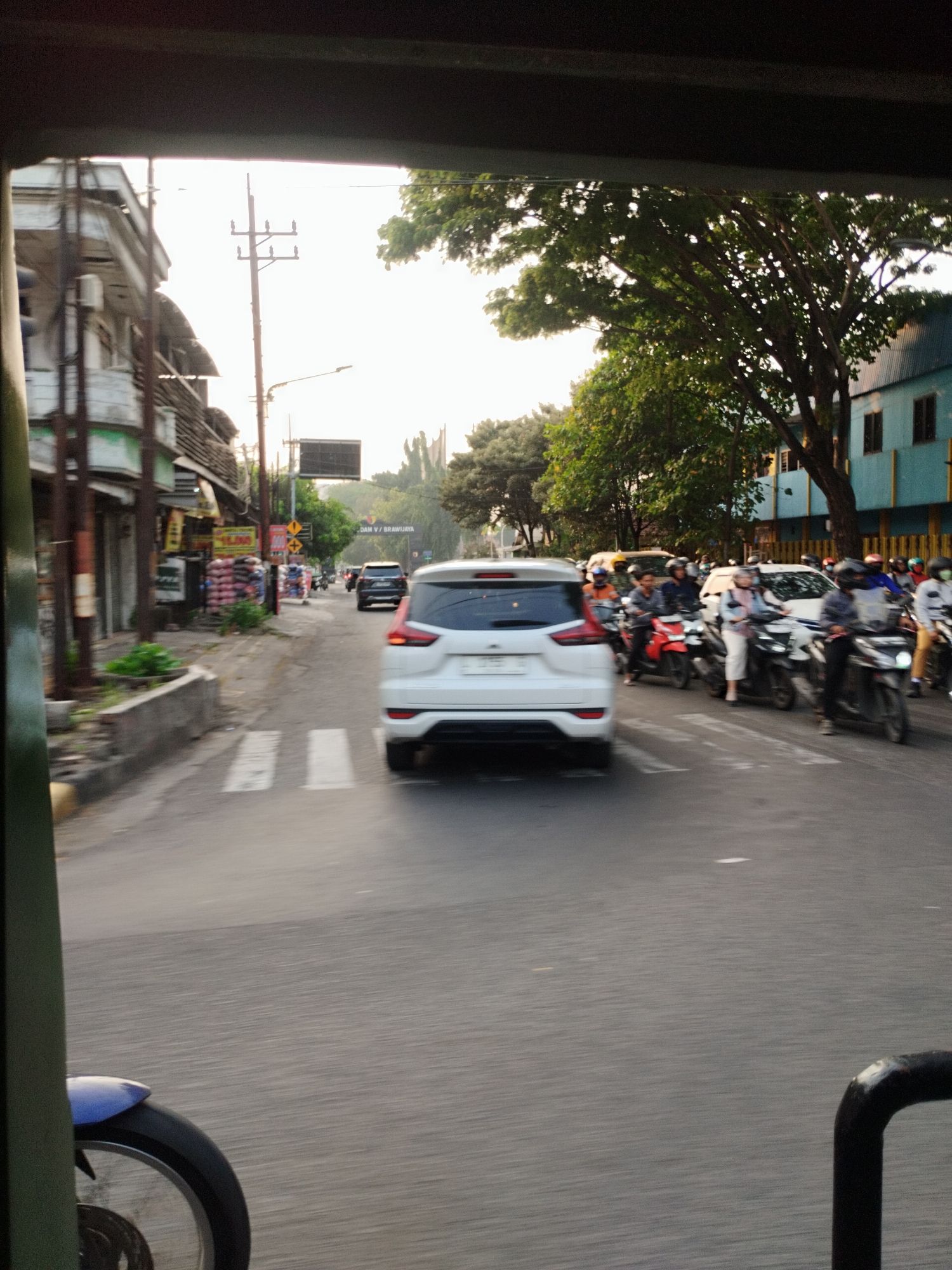 The image depicts a street scene in Surabaya, Indonesia, likely during daylight hours given the bright sky. The perspective is from inside a vehicle, possibly a becak or similar, looking forward. In the foreground, a part of the vehicle's frame and wheel are visible on the left side. The street is paved and shows white pedestrian crossing lines.
Several vehicles are present on the road. A white SUV is the most prominent, seen from its rear, driving away from the viewer. Further down the road, other cars and numerous motorcycles are visible, many with riders wearing helmets. Some motorcycles are parked along the side of the road.
The street is lined with buildings on the left, which appear to be shops or businesses with awnings. There are signs with text and some displays of merchandise. On the right side of the street, trees with dense green foliage overhang the road, and a few buildings are visible, one of which is light blue. Power lines and poles are also present along the street. The overall impression is a busy urban street with moderate traffic. The image is slightly blurred, suggesting movement.