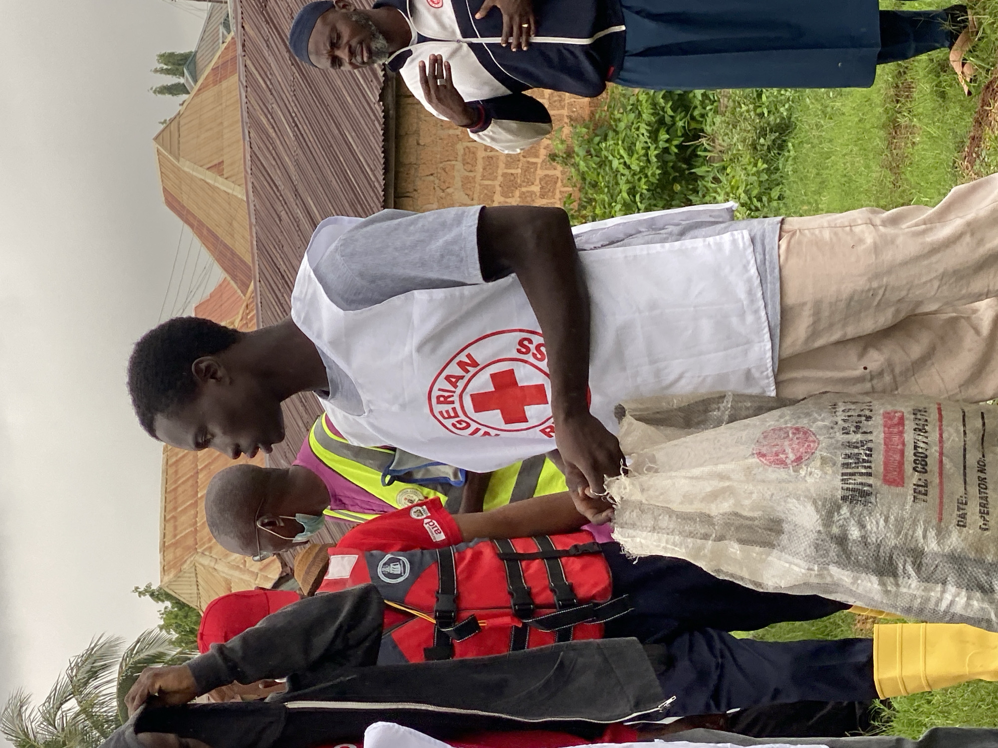 In Kawo, Nigeria, a group of people are gathered outdoors. In the foreground, a young man wears a white vest with a red cross emblem and "NIGERIAN RED CROSS" written on it. He is holding a large, transparent plastic bag filled with dark material. Next to him, another person in a red life vest with black straps and a white collar is visible. To the right, a man in a blue and white jacket and a dark blue skirt stands with his hands clasped. Behind them, several other individuals are present, some wearing brightly colored vests. The background shows buildings with corrugated metal roofs and some greenery, suggesting a residential or community area. The sky is overcast.