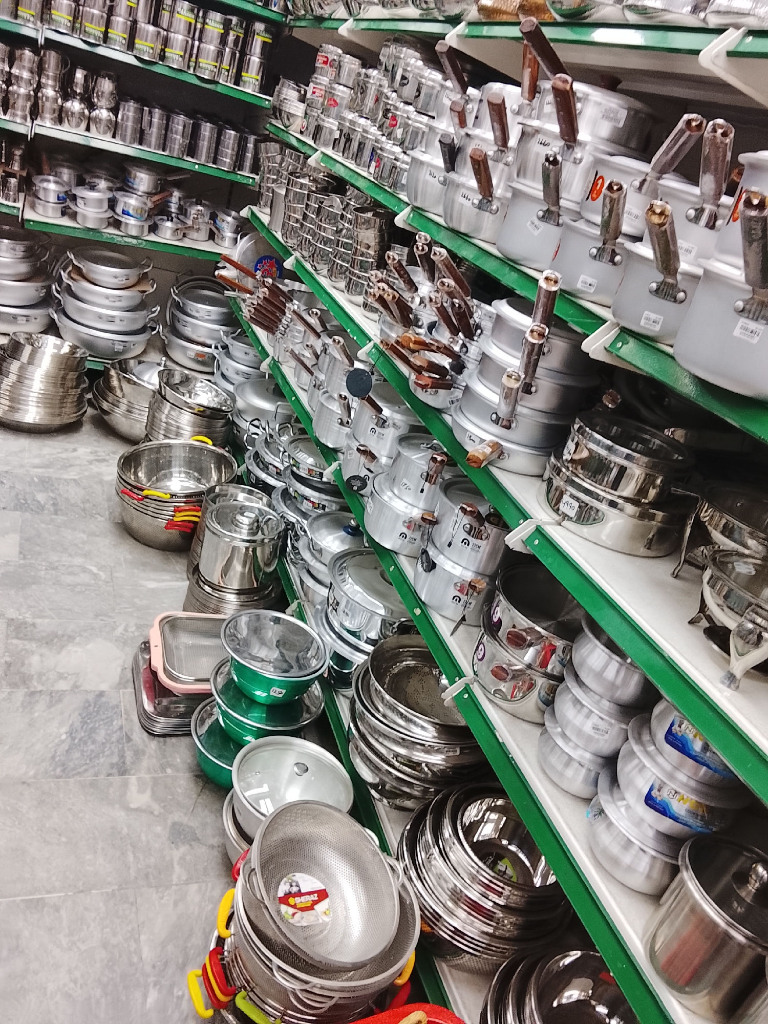 The image shows a retail store in Gulgasht Colony, Pakistan, specializing in cookware. Shelves lined with stainless steel and aluminum pots, pans, and bowls dominate the scene. The merchandise is densely packed, ranging from large cooking pots to smaller saucepans and colanders, reflecting a variety of kitchenware options. The shelves themselves are constructed with green-edged supports. The floor is a light gray, possibly tiled or finished concrete. No people are visible in the frame.