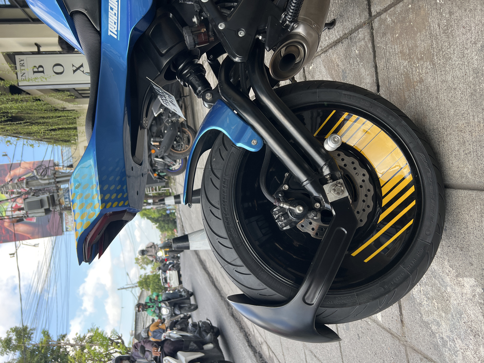 The image shows the front wheel and part of the front body of a blue motorcycle. The wheel is black with yellow stripes. The tire brand is Michelin Pilot Street Radial. In the background, there is street traffic including other motorbikes, and a sign that says "ENTRY BOX". Based on the street scene and sign, this photograph was likely taken in North Kuta, Indonesia.