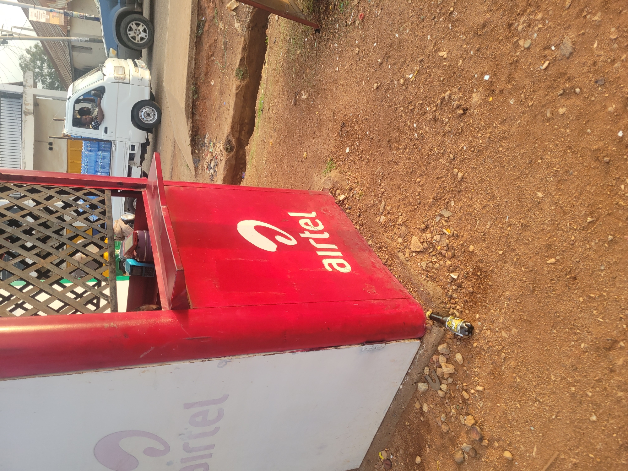 The image depicts an Airtel kiosk on a street in an unknown city. The kiosk, painted red and white with the Airtel logo, is situated on a dirt and gravel area. A discarded beer bottle rests near the kiosk's base. A white truck carrying water bottles is visible in the background, along with a blue vehicle and some buildings. Two people are visible inside the truck.