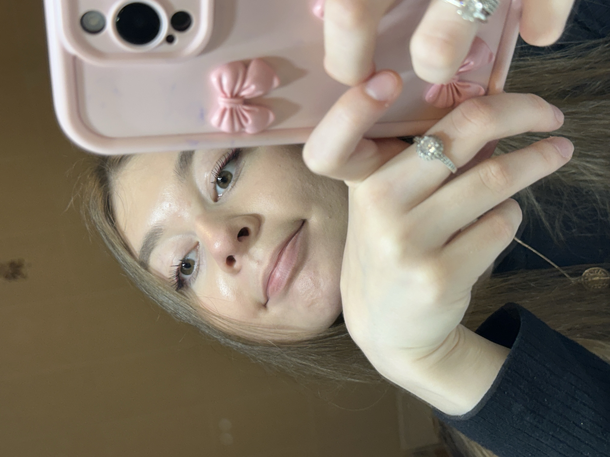 The image is a close-up, tilted selfie of a young woman looking directly into the camera, holding a pink smartphone. Her face is positioned at an angle, with her head slightly to the left and her gaze directed upwards.
The subject is a woman with light skin, brown hair, and defined eyebrows. Her eyes are brown, and she has visible dark eyelashes, suggesting mascara. Her lips are a natural pink hue, and she has a subtle, soft smile. She wears a black long-sleeved top with a ribbed texture, visible around her right arm. Around her neck, a thin chain necklace with a small, round, possibly metallic pendant is visible.
She is holding a pink smartphone, which covers the upper portion of her head. The phone case is adorned with two distinct three-dimensional pink bow embellishments. The camera lenses of the phone are visible at the top of the case. Her right hand, from the viewer's perspective, is holding the phone, showcasing a silver-toned ring with a prominent clear or light-colored stone on her index finger. Another silver-toned ring with multiple stones is visible on a finger of her left hand, which is partially obscured. Her fingernails are short and natural.
The background is uniformly blurry and appears to be a plain, warm brown or beige wall, indicating an indoor setting. There is no discernible text or branding visible on any of the objects in the image. The overall mood is casual and direct.