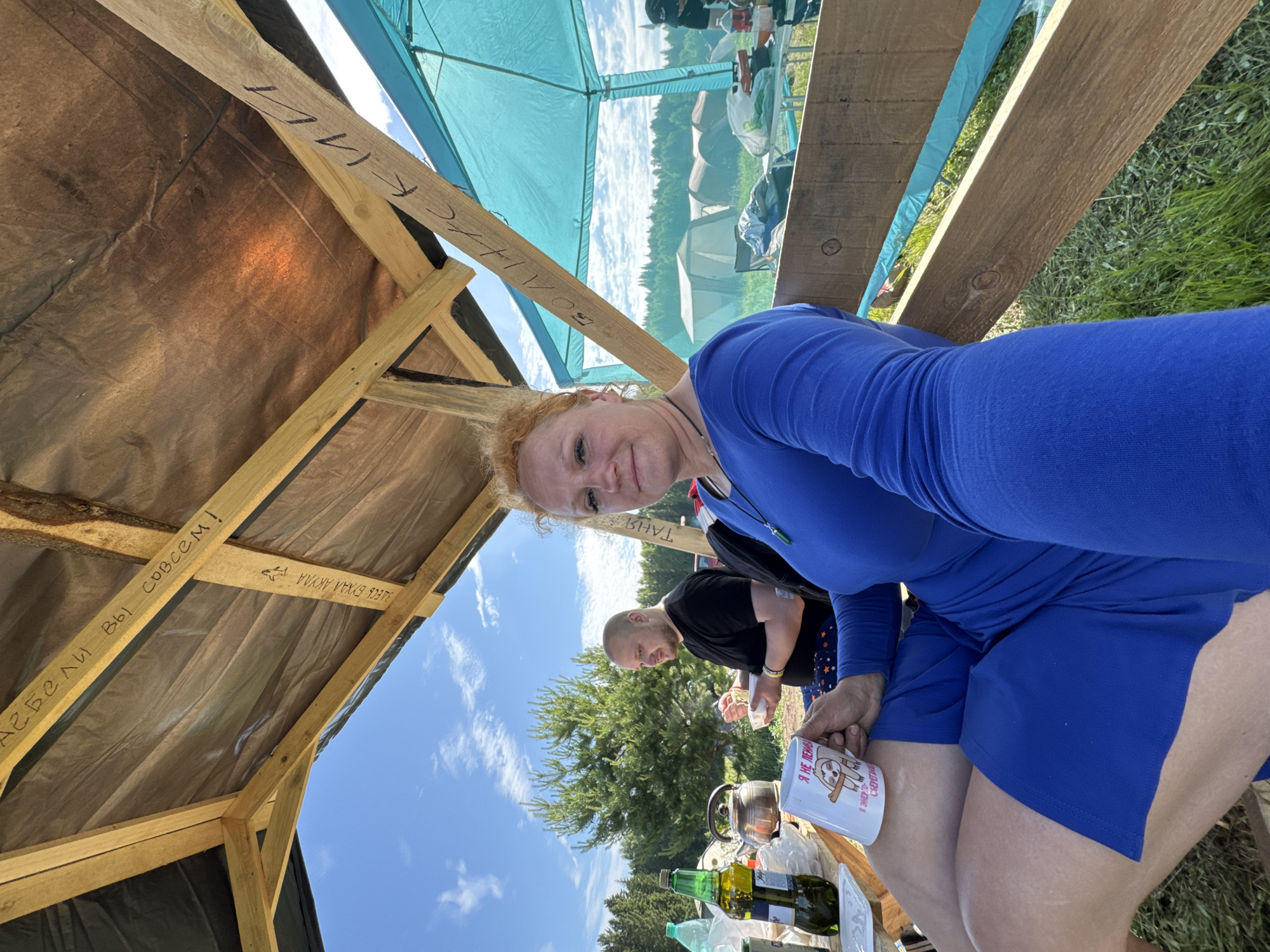 This outdoor image depicts a woman taking a selfie within a temporary wooden and tarp structure, featuring two individuals and a campsite environment. The location is Большая Кивара, Russia.
In the immediate foreground, a woman with reddish-blonde curly hair and fair skin is visible from the chest down, positioned on the right side of the frame. She wears a long-sleeved royal blue garment, possibly a dress or top paired with shorts. She holds a white mug in her right hand, which features an illustration of a sloth and Russian text reading "Я НЕ ЛЕНИВАЯ, Я ЭНЕРГИЮ СБЕРЕГАЮ" (I'm not lazy, I'm saving energy). A green pendant hangs from her neck. Her gaze is directed towards the camera, suggesting she is capturing the image.
To her left, a man with a bald head or very short dark hair, wearing a black t-shirt and patterned blue shorts, is seated. He holds a white bowl, appearing to be eating. Various items are on a wooden surface in front of them, including a green glass bottle with a white label, a dark metallic kettle on a small burner, and other small containers.
The setting is defined by a rustic wooden frame with a dark tarp serving as a roof, providing shade. Visible handwritten inscriptions on the wooden beams include "ЕБЕЛИ ВЫ СОВСЕМ!" (a rude Russian phrase meaning "You're completely fucked/screwed!"), "ЗДЕСЬ БУХАЛ АЙКЫЛ" (Aikyl was drinking here), "БОЛТЫСК" (an unclear word, possibly a name or slang), and "ТАНЯ" (Tanya).
Beyond the structure, a clear blue sky with scattered white clouds is visible. In the background, a dense coniferous forest stretches across the horizon. Several light-colored tents are pitched in a grassy field, indicating a campsite. To the far right, a light blue canopy tent is also visible. The ground under the wooden structure is covered in green grass.