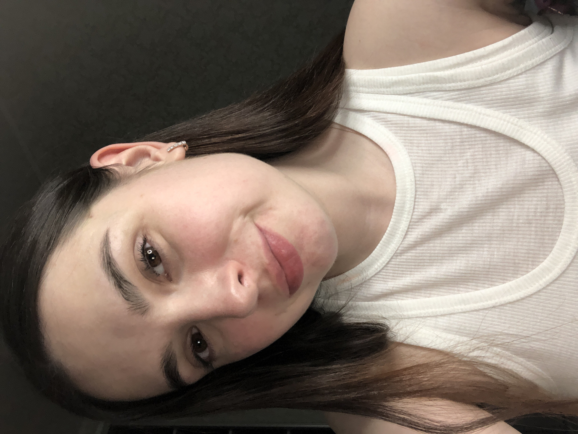 A woman with brown hair is wearing a white ribbed tank top. She has brown eyes, fair skin, and a silver earring in her left ear. The background is a dark, possibly gray or black, color. No information about the location is available, as the image does not provide any environmental cues, nor can it be assumed.