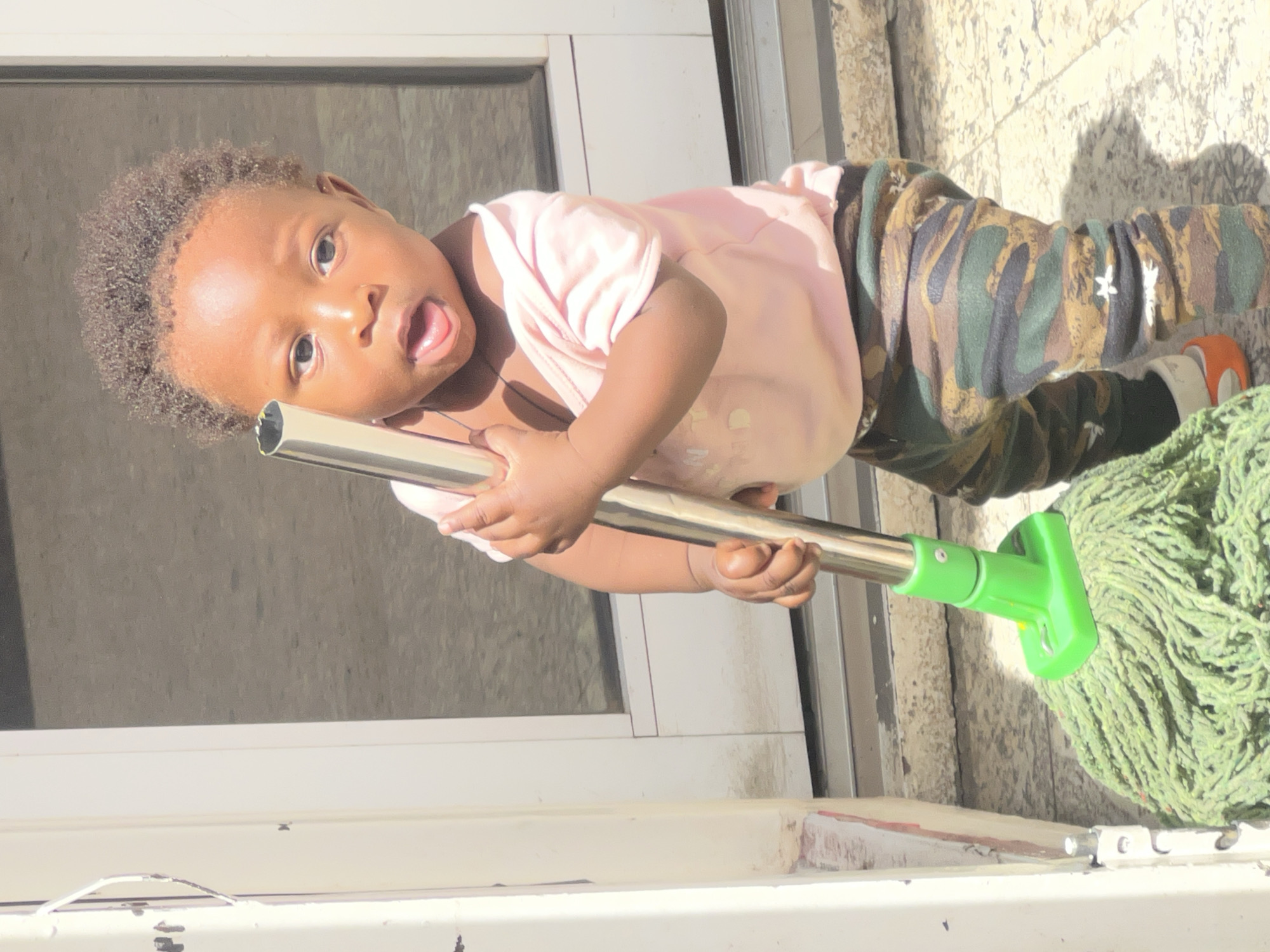 A young child, possibly a toddler, is holding a mop outside a building in Gura Top, Nigeria. The child is wearing a pink shirt and camouflage pants, and appears to be playing with the mop. The background shows a window and the exterior of a building. The child has the mop at an angle pointing from left to right and downward. The child's mouth is open.