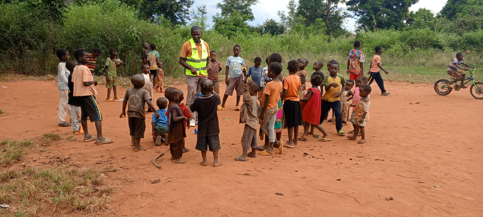 In Ngalula, Democratic Republic of the Congo, a group of approximately 25 children is gathered in a dirt clearing near vegetation. An adult male wearing a yellow reflective vest stands amongst the children. A boy rides a bicycle in the background.