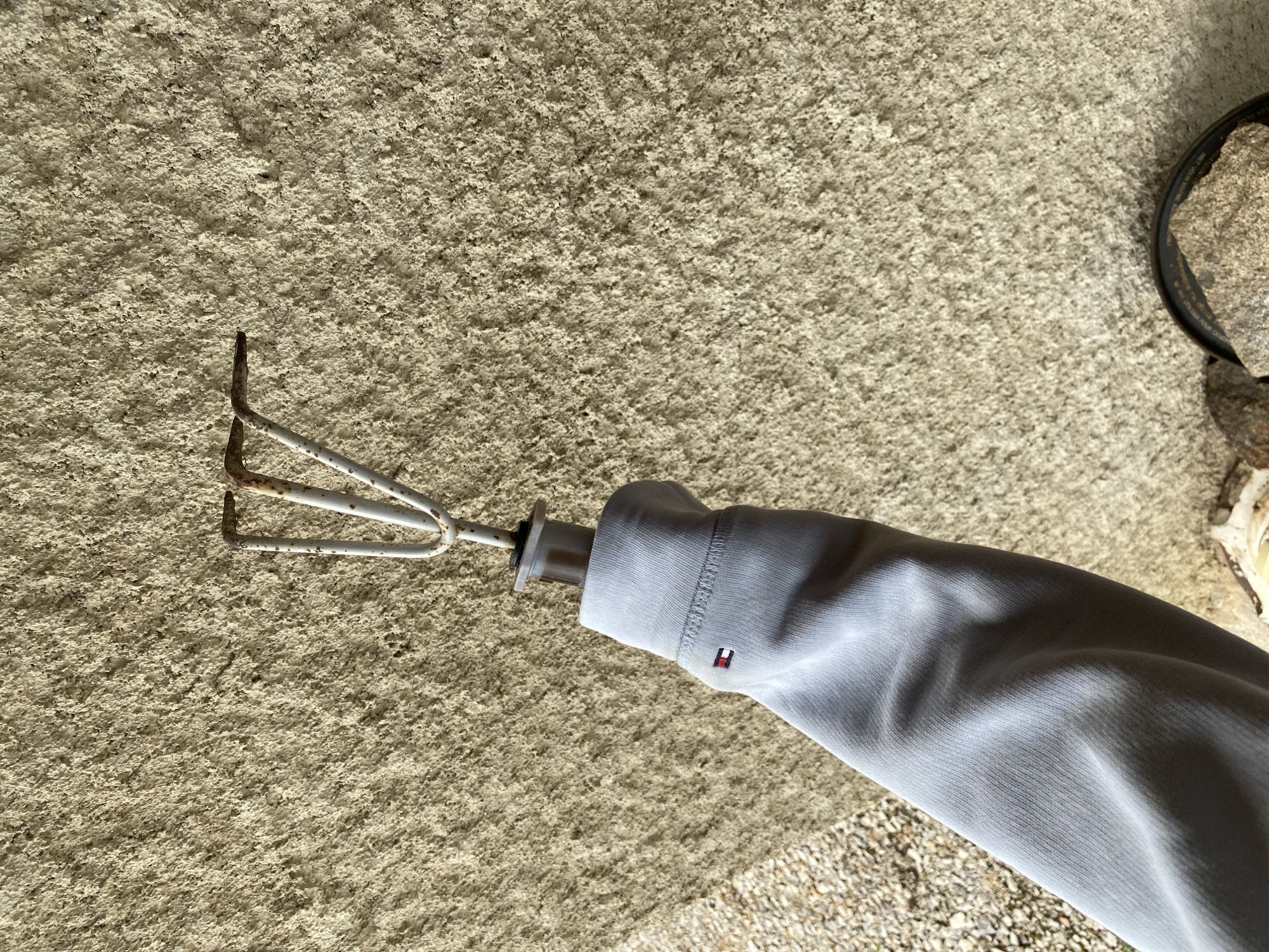 The image captures a close-up of a person's arm, clad in a light grey long-sleeved garment, holding a small metal gardening tool. The tool is a hand cultivator or rake with three curved prongs, painted white or light grey, showing signs of minor rust at the tips. A white plastic component connects the tool's head to its handle, which is mostly concealed by the sleeve. On the cuff of the garment, a small embroidered logo is visible, featuring a rectangular design with red, white, and blue sections, identifiable as the Tommy Hilfiger brand emblem.
The background consists of a rough, granular, light-colored surface, possibly textured concrete, stucco, or a coarse-grained pathway. Towards the bottom right corner, a patch of smaller, dark grey stones or gravel is evident. On the far right edge of the frame, a partial view of a dark, circular object, possibly a pot or a drain, can be seen.
The activity depicted is the holding of the gardening tool, suggesting either preparation for gardening work or a momentary pause during it. The scene is brightly lit, indicating it is daytime. The location context is Courpière, France.