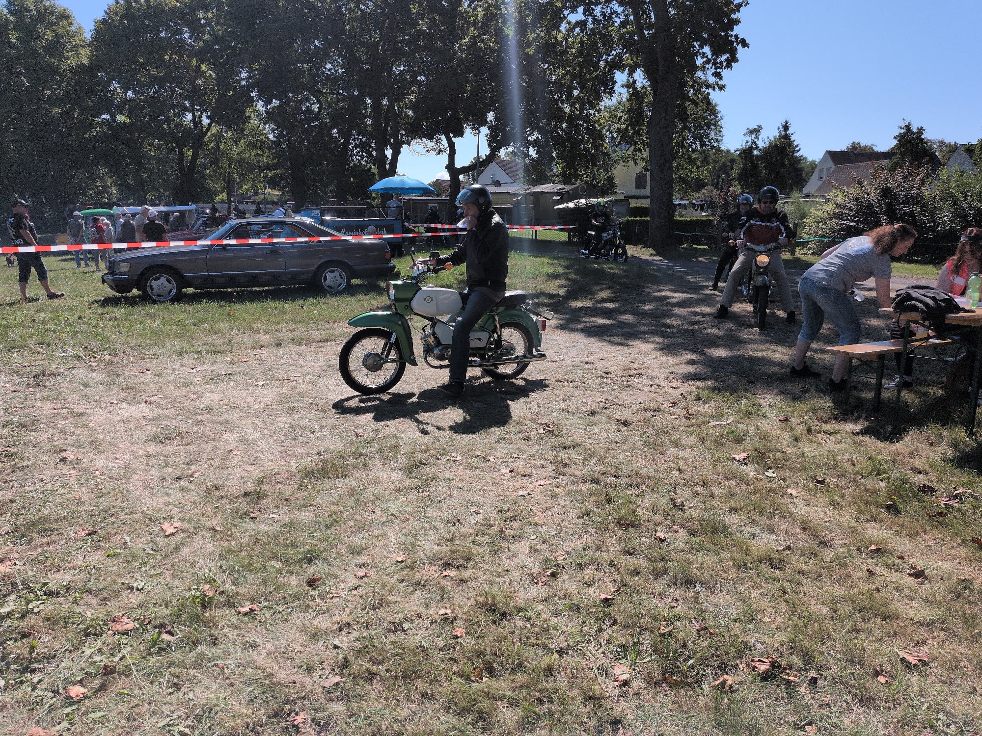The image depicts an outdoor event in Hohenferchesar, Germany, likely a gathering of classic cars and motorcycles. In the foreground, a person is riding a vintage green and white motorcycle. To the left, a classic grey Mercedes-Benz sedan is parked on a grassy area, cordoned off by red and white tape. Several people are standing and observing the vehicles. In the background, more vehicles, including motorcycles, are visible, along with trees and buildings. The weather appears to be sunny and clear.