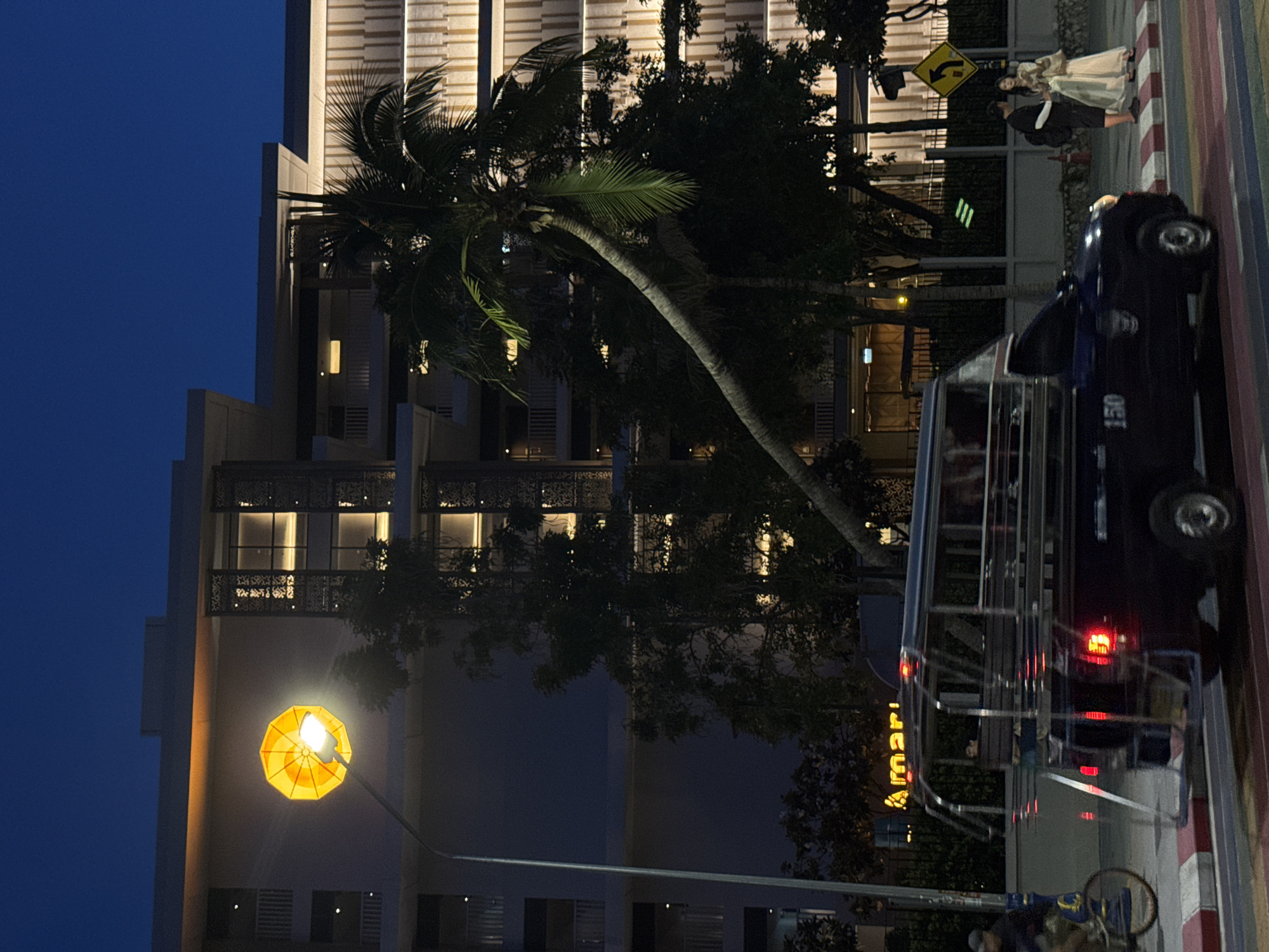 The image captures a street scene in Pattaya City, Thailand, at dusk. A distinctive street lamp with an umbrella-shaped, illuminated shade stands on the left, casting a warm glow. In the background, a modern building with illuminated windows and decorative elements is visible. A palm tree leans across the scene, its fronds silhouetted against the twilight sky. A dark-colored, open-back vehicle, possibly a songthaew, is driving on the road, its brake lights illuminated. To the left of the vehicle, a person on a bicycle is in motion. On the right side of the road, a couple, a man in a dark shirt and a woman in a light-colored dress, stand together. A yellow traffic sign with a black arrow indicating a turn is also present on the right. The street is marked with a rainbow-colored crosswalk. The overall atmosphere suggests a vibrant urban environment transitioning into night.