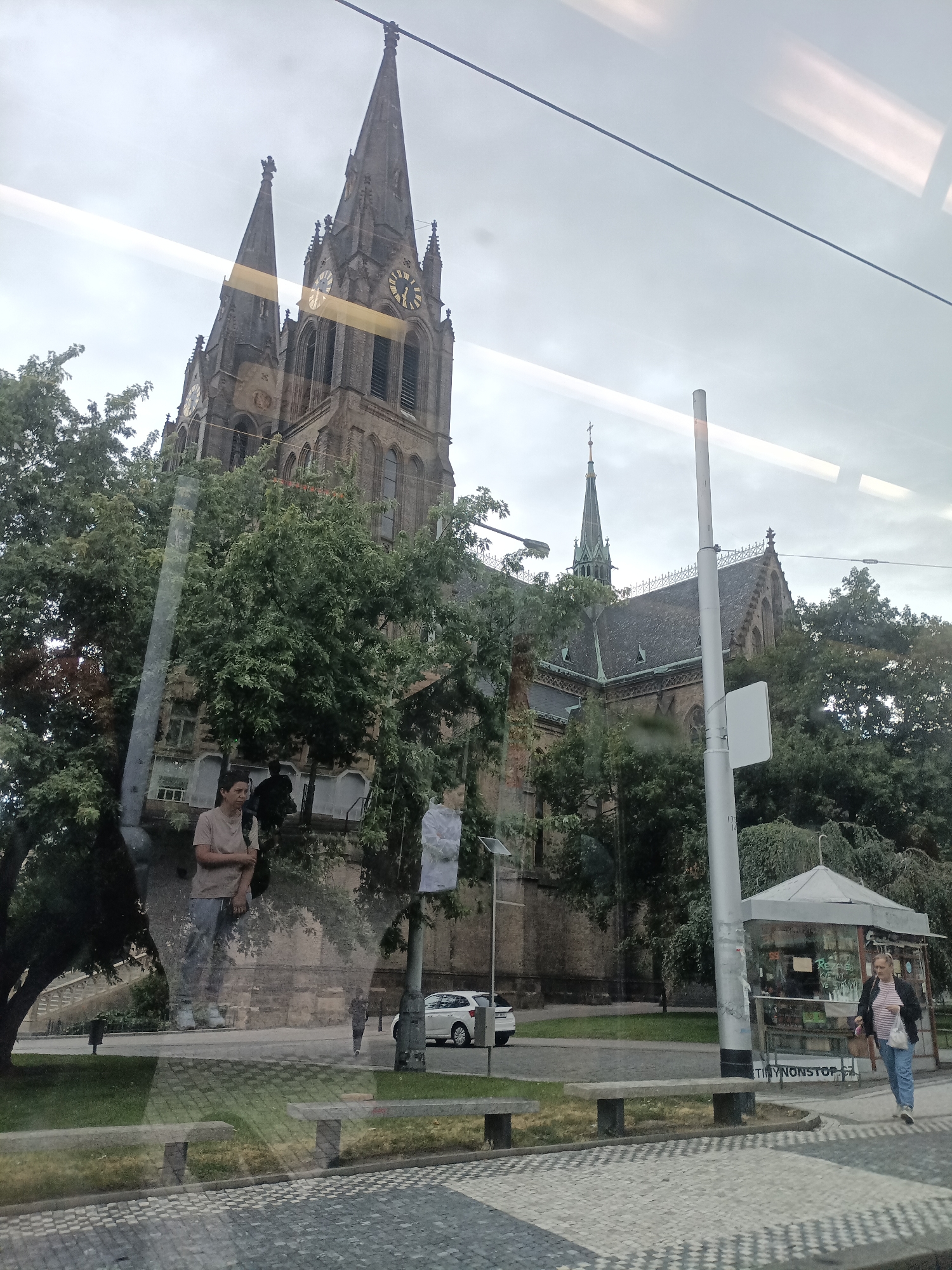 The image shows a street scene in Prague, Czechia, with a large church and trees in the background. The church has two tall spires, one with a clock face. In the foreground, there are benches, a sidewalk made of cobblestones, and a newspaper kiosk. A woman is walking towards the right, carrying a bag. The scene is captured through a window, as evidenced by reflections on the glass.