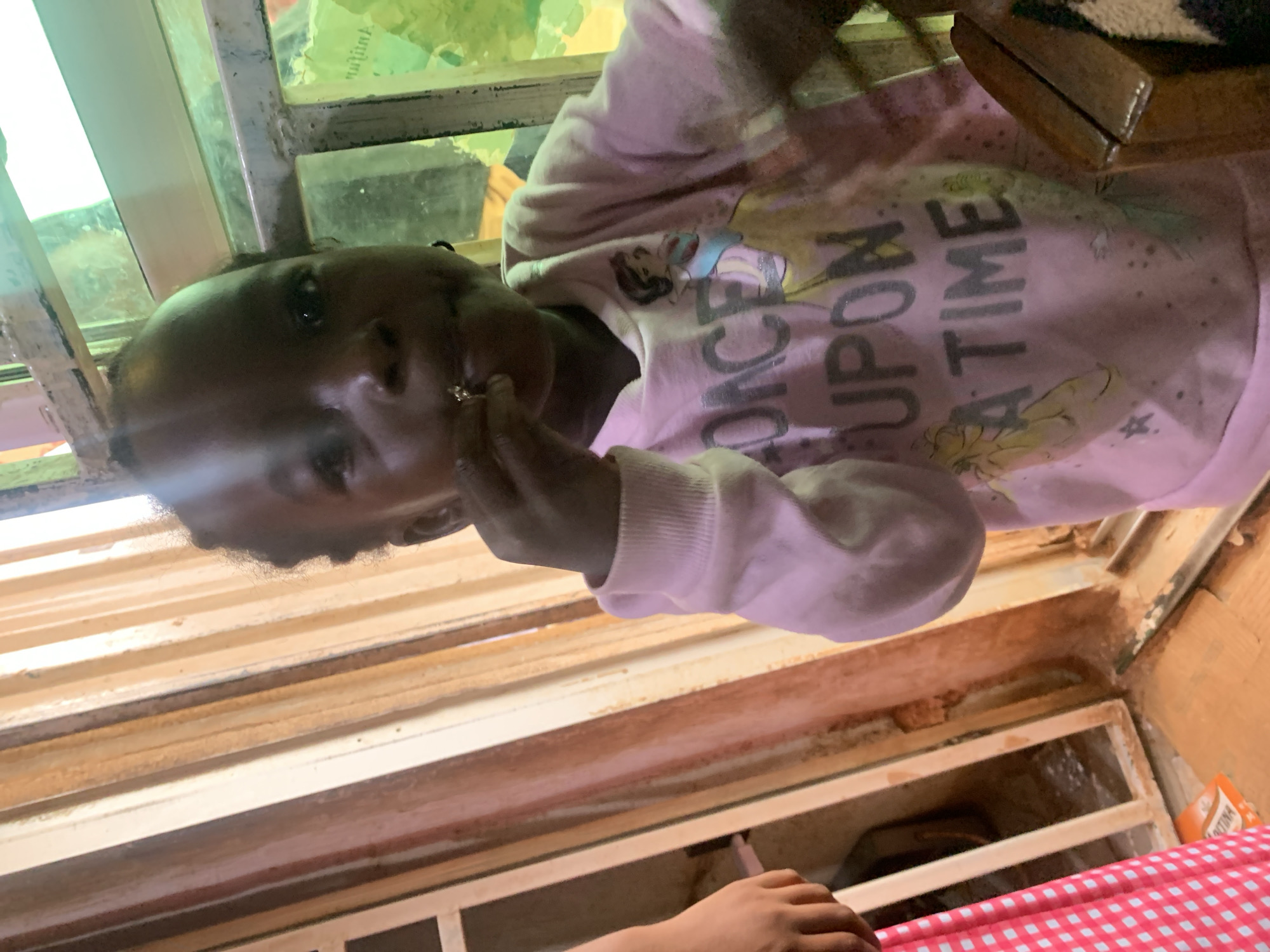 A young child, with dark skin and hair, is shown from the chest up, looking at the camera and bringing a small object to their mouth with their right hand. The child wears a light pink sweatshirt with a Disney princess graphic and the text "ONCE UPON A TIME" in grey lettering. The child is positioned indoors, near a window with colorful panes and a rough, textured frame. To the left of the child, another person's arm and hand are visible, wearing a pink and white checkered garment. The background features vertical wooden slats and a metal grate. The lighting is dim, with a greenish hue from the window. This scene is likely in Sabon Tasha, Nigeria, a district in Kaduna State.