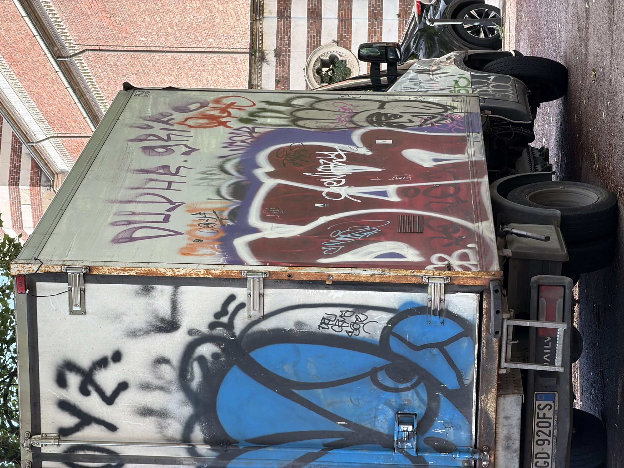 The image shows the rear of a graffiti-covered truck, parked in the city of Unknown, Unknown. The truck has a white body, with a blue and black graffiti design on the lower half of the back door, and a red, purple, and orange design on the upper half. The license plate is visible and reads "CD 920FS". The vehicle is parked on a street, with a building in the background constructed of red and white bricks. A black car is parked in front of the truck.