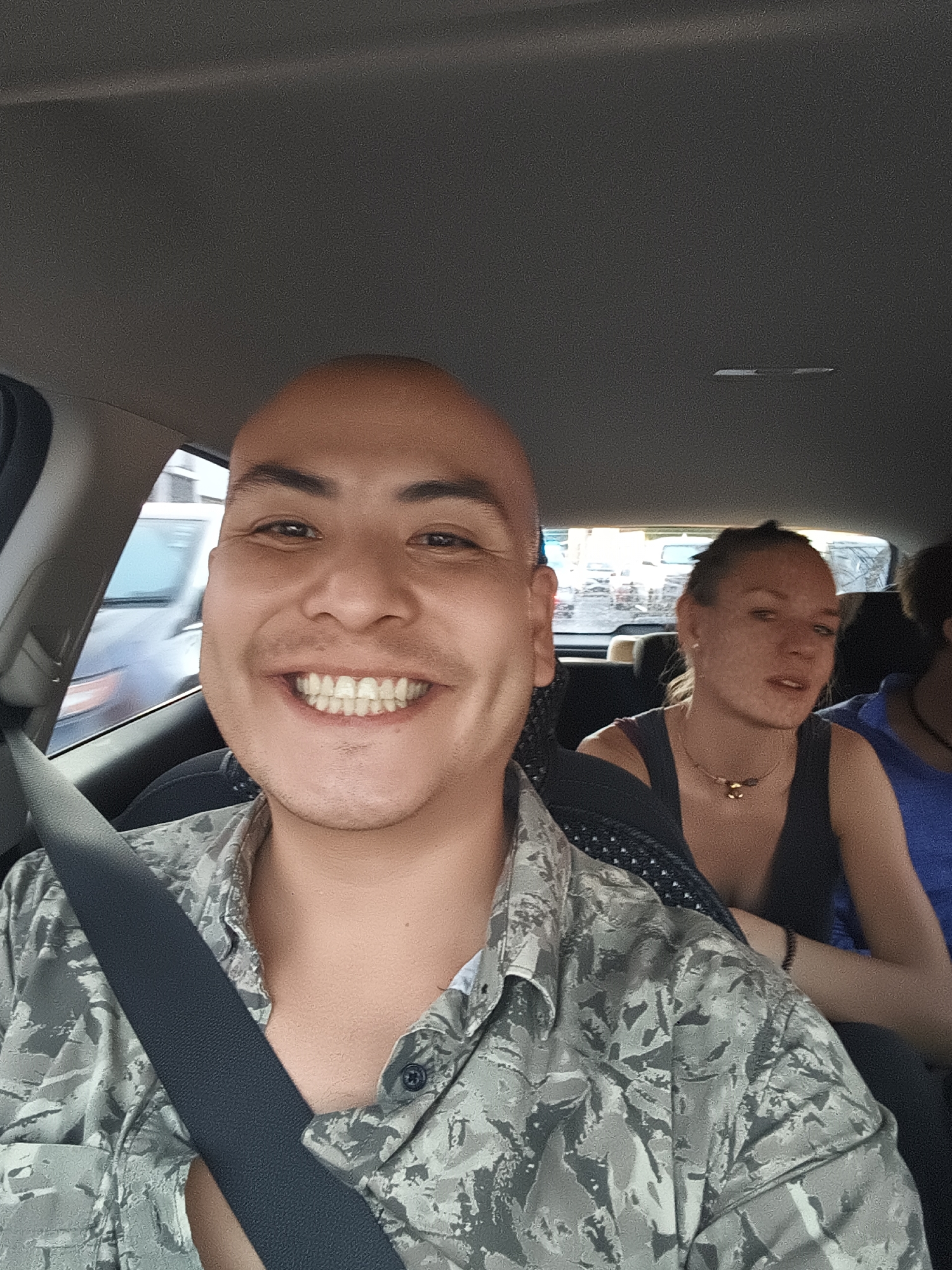 The image is a selfie taken from the front passenger seat inside a vehicle, likely a car. The person taking the selfie is a bald man wearing a camouflage-patterned shirt and a seatbelt. He is smiling broadly at the camera.
In the back seat, two other individuals are partially visible. The person directly behind the selfie-taker is a woman with her hair pulled back, wearing a black tank top and a necklace. The person behind her, partially obscured, is wearing a blue shirt.
Through the car windows, a city street scene is visible. The location is identified as the city of Querétaro, Mexico, based on general architectural styles and vehicle types seen in the background, along with the fact that it appears to be a very old colonial city. Several vehicles and buildings are visible through the windows, indicating the car is on a city street with traffic. The overall impression is of a daytime outing in an urban setting.