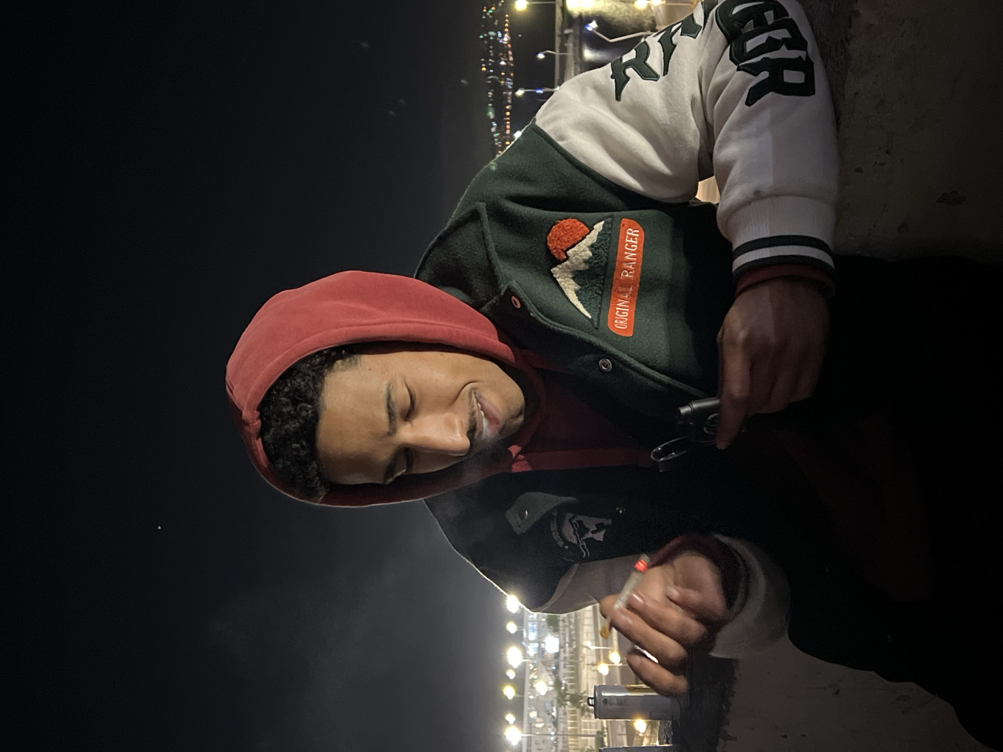 A male individual is depicted outdoors at night in Tanger, Morocco, positioned with his upper body visible. He has dark, curly hair and a mustache, with his eyes closed or squinted as he exhales smoke. He wears a red hooded sweatshirt underneath a dark green and white varsity jacket. The jacket features an embroidered patch on the left chest with a red sun, white mountains, and the text "ORIGINAL RANGER". In his right hand, he holds a lit cigarette with a visible ember. His left hand holds a dark, cylindrical object, possibly a lighter, attached to a silver carabiner clip. He leans on or is seated behind a concrete barrier. The background consists of numerous blurred artificial lights from a cityscape or waterfront, indicative of an urban environment at night.