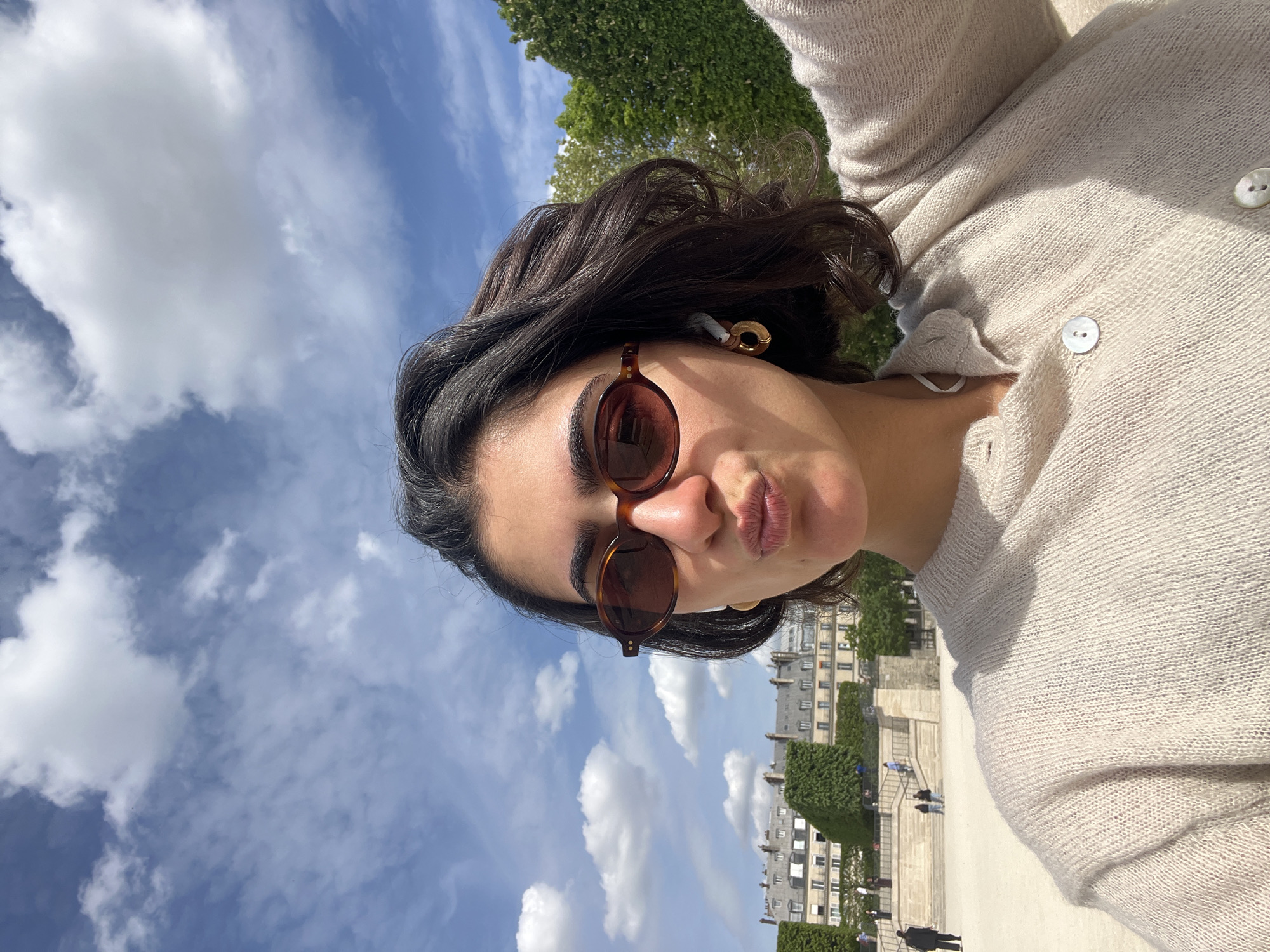 A bright, outdoor selfie captures a young woman in Paris, France, under a clear blue sky with scattered white clouds. The woman, with dark, wavy hair, is in the foreground, wearing tortoiseshell-framed sunglasses and a light beige or off-white knitted cardigan with white buttons. She poses playfully with her lips slightly pursed. A gold-toned earring is visible on her right ear, along with a white wireless earbud.
The background reveals a sunny day over what appears to be a public park or garden. Green trees and manicured bushes frame stone steps and pathways, with several people walking in the distance. Beyond the immediate park setting, classic Parisian architecture with light-colored buildings and multiple windows confirms the location as Paris. The bright daylight suggests it is midday or early afternoon. No visible text is present in the image.
