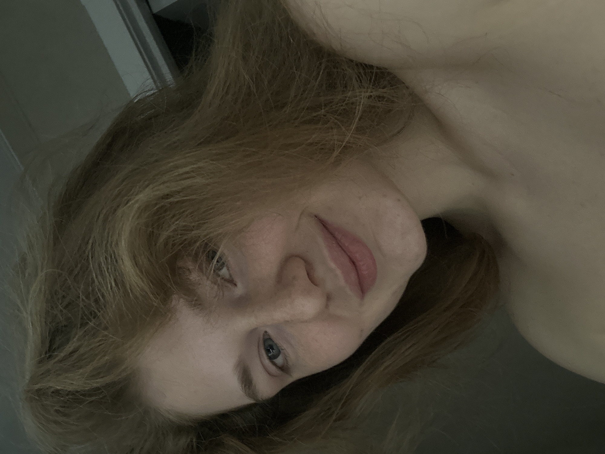 A close-up, angled headshot of a fair-skinned person with long, light brown hair. The person's face is slightly tilted, with a subtle smile. Their eyes are blue. The background is indistinct but appears to be an interior space, with a door frame visible in the upper left. There is insufficient information to locate the image in Kazan, Russia.